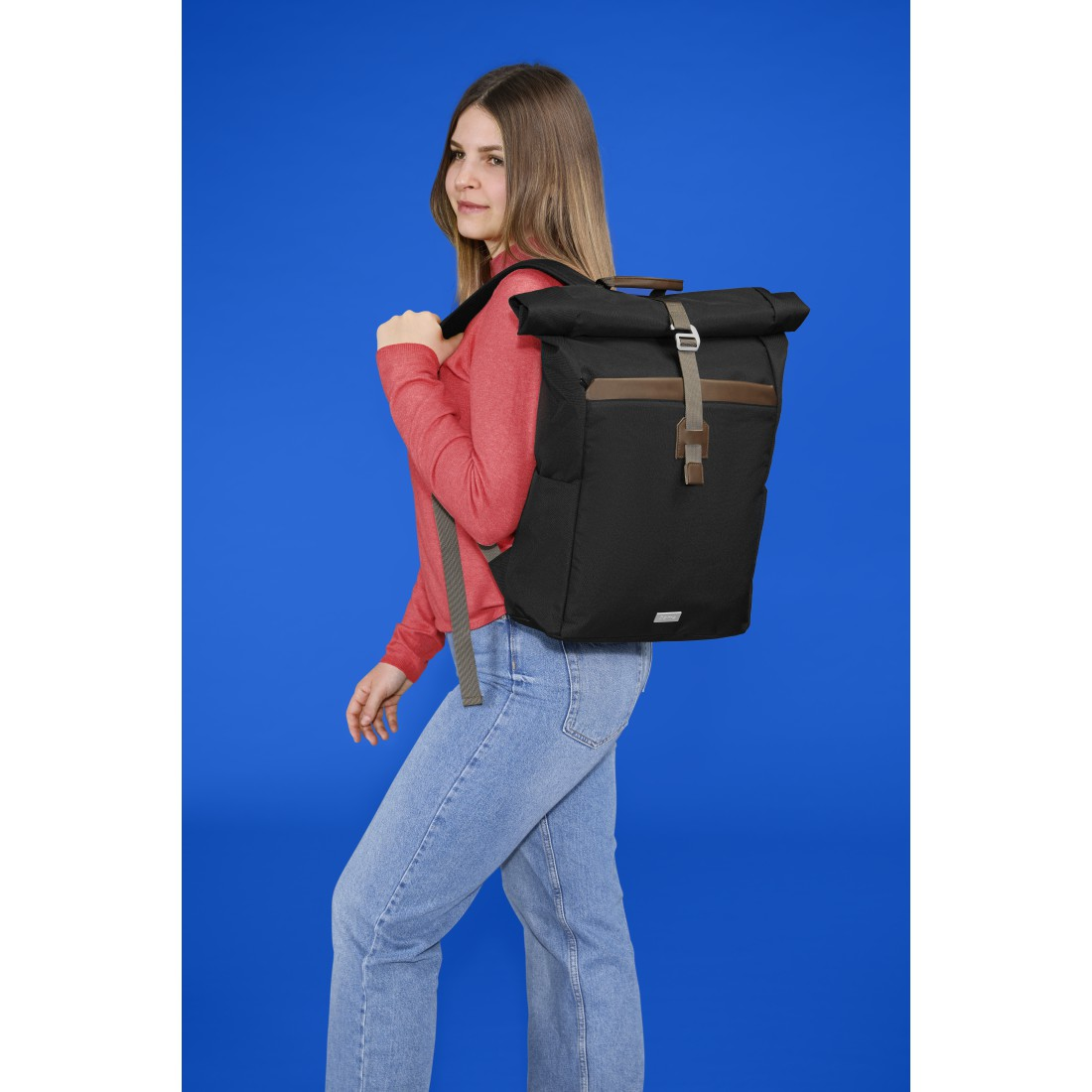 Frau mit rotem Pullover und Jeans, mit schwarzem Rucksack vor blauem Hintergrund.