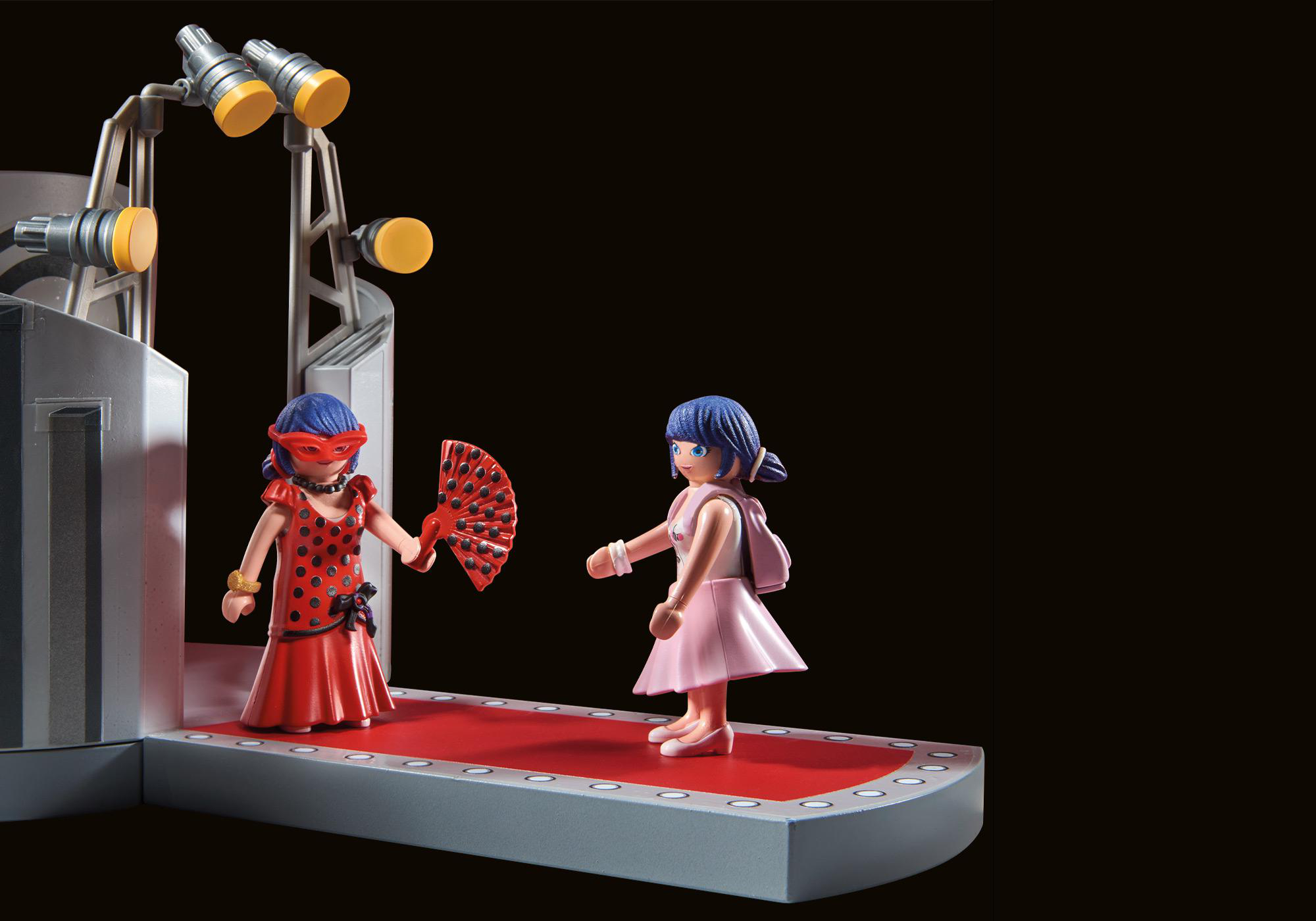 Zwei Figuren auf einer Bühne. Eine trägt ein rotes Kleid und eine Maske, die andere ein rosa Kleid.