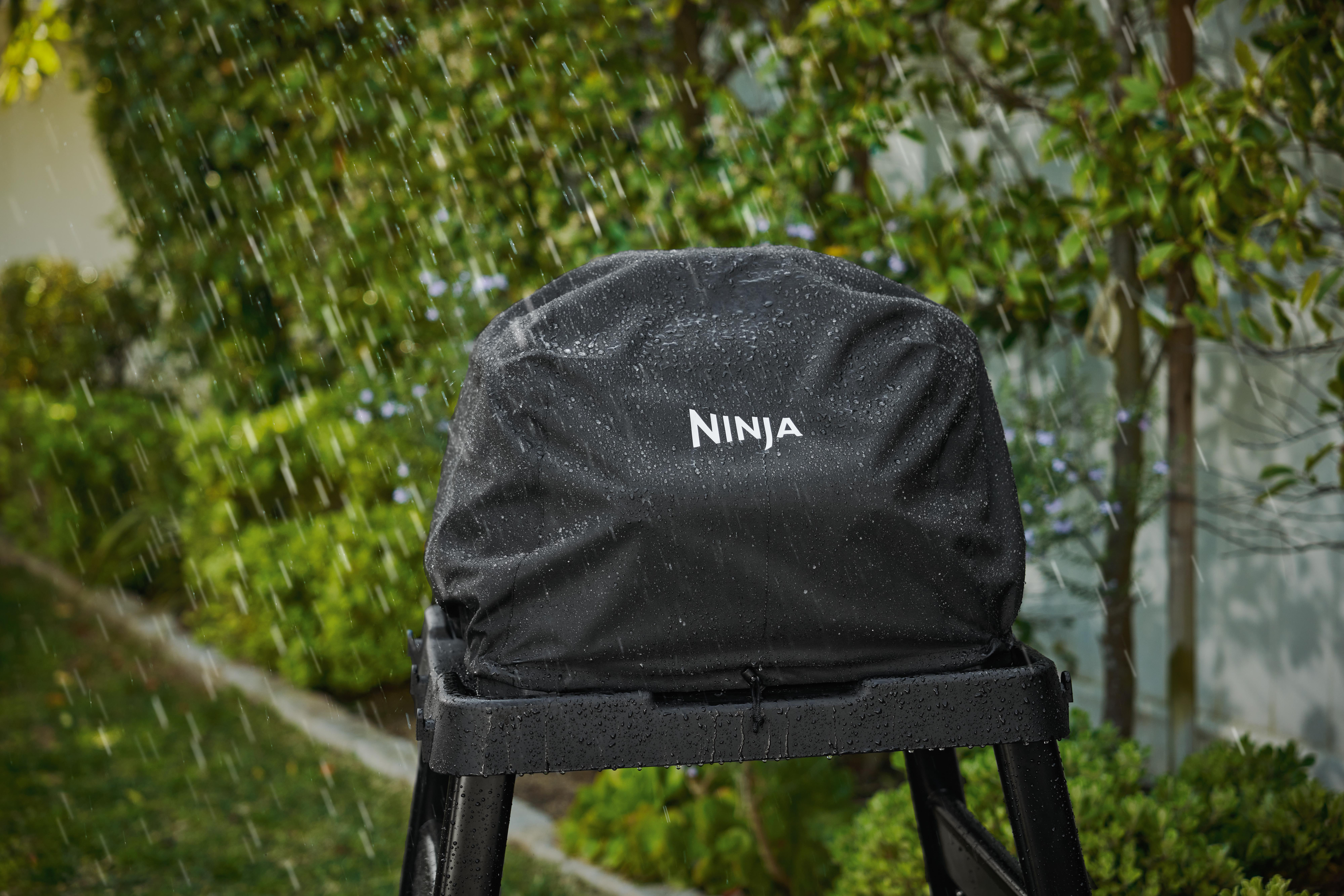 Schwarzer Ninja-Grill, bedeckt mit einer schwarzen Abdeckung. Regen fällt. Grüner Hintergrund von Bäumen.