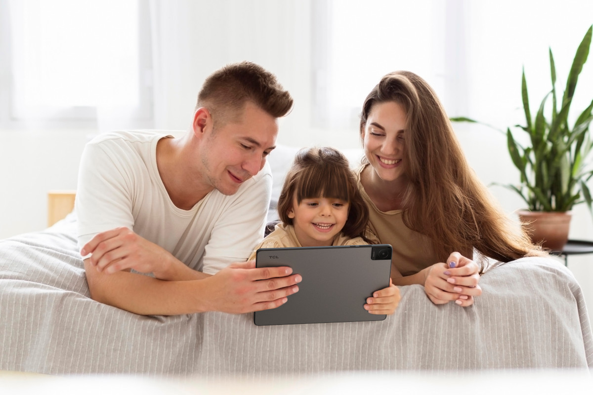 Rodzina leży na łóżku, patrząc na tablet. Uśmiechają się i wchodzą w interakcję z urządzeniem.