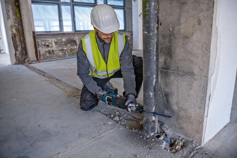 Egy építőmunkás védőfelszerelésben fűrészt használ beton vágásához egy építkezésen.