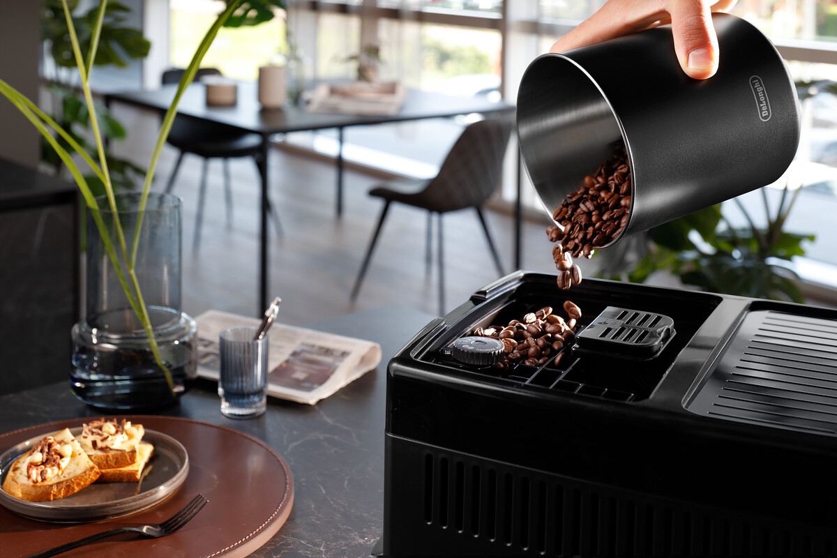 Ziarna kawy są wlewane do czarnego ekspresu do kawy. W tle stół.