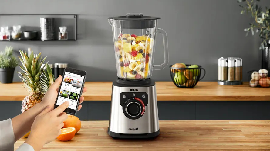 Blender z owocami, osoba korzystająca z telefonu i drewniany blat z akcesoriami kuchennymi.