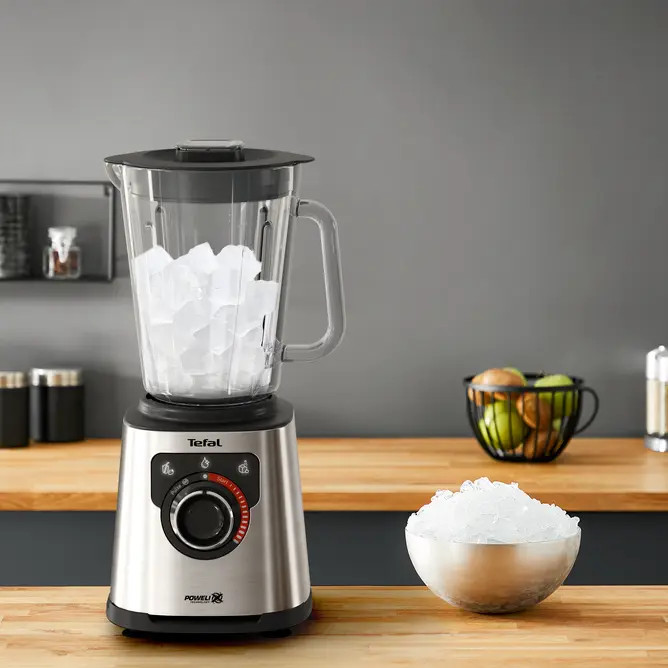Blender Tefal z lodem. Blender stoi na stole z miską lodu po prawej stronie i ciemnym tle.