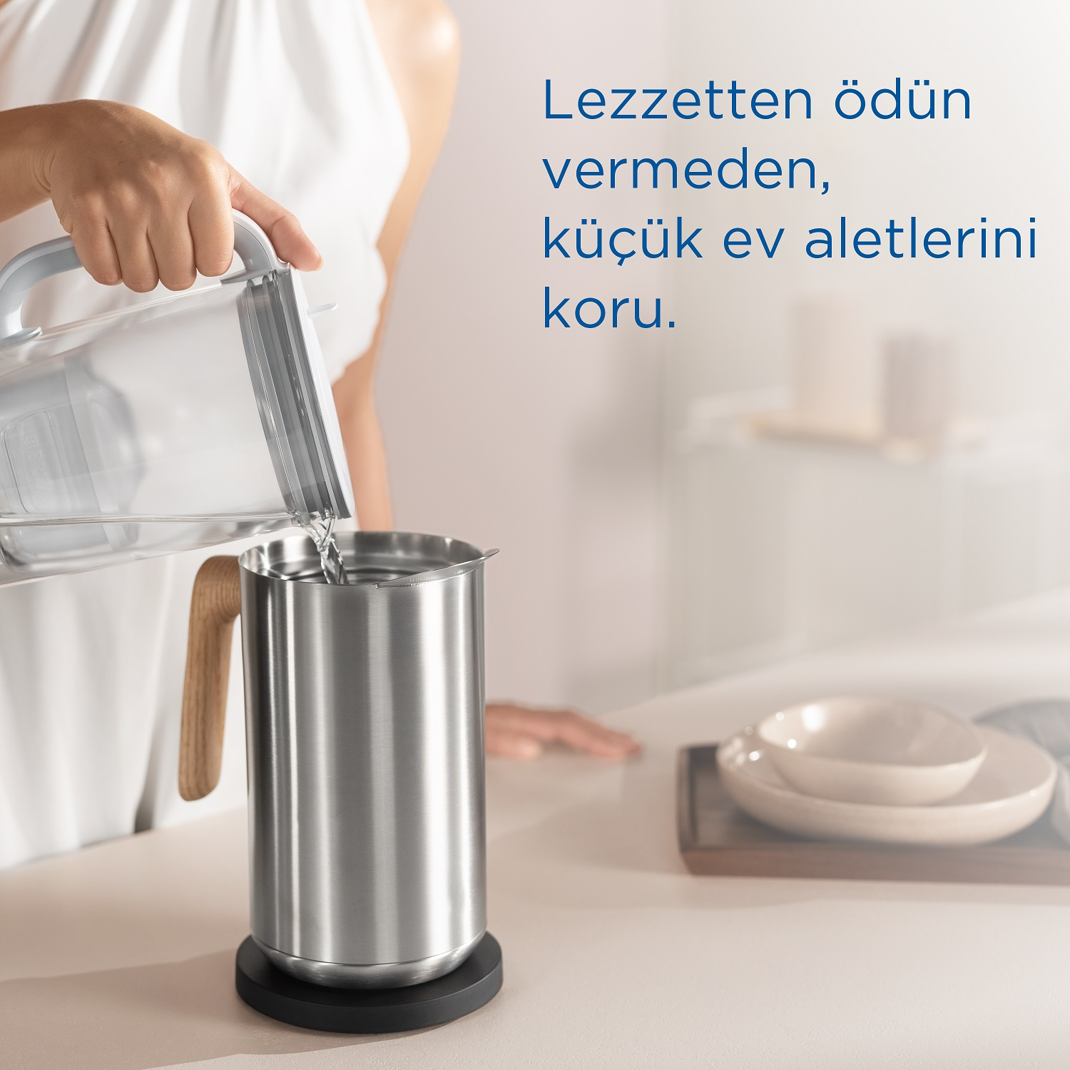 Bir kadın, beyaz bir yüzey üzerinde ahşap saplı paslanmaz çelik bir sürahiye su döküyor.