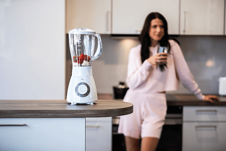 Un blender blanc avec des ingrédients et une femme tenant un verre dans une cuisine.