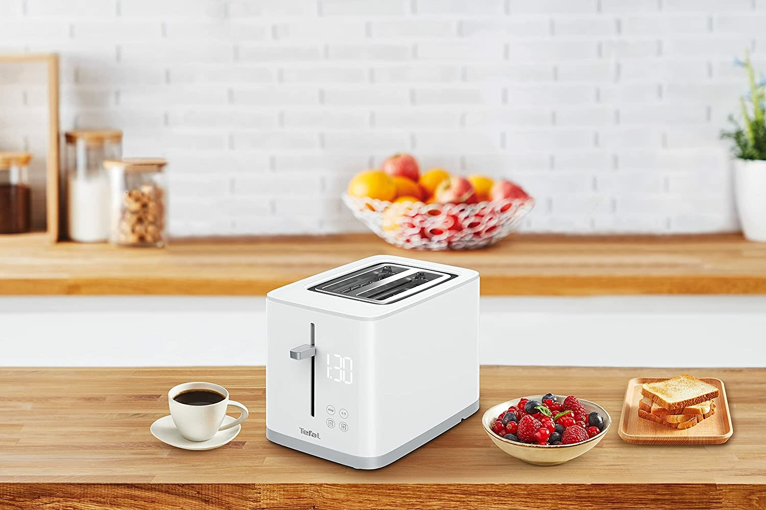 Biały toster Tefal z wyświetlaczem cyfrowym. Obok chleb, jagody i kawa.