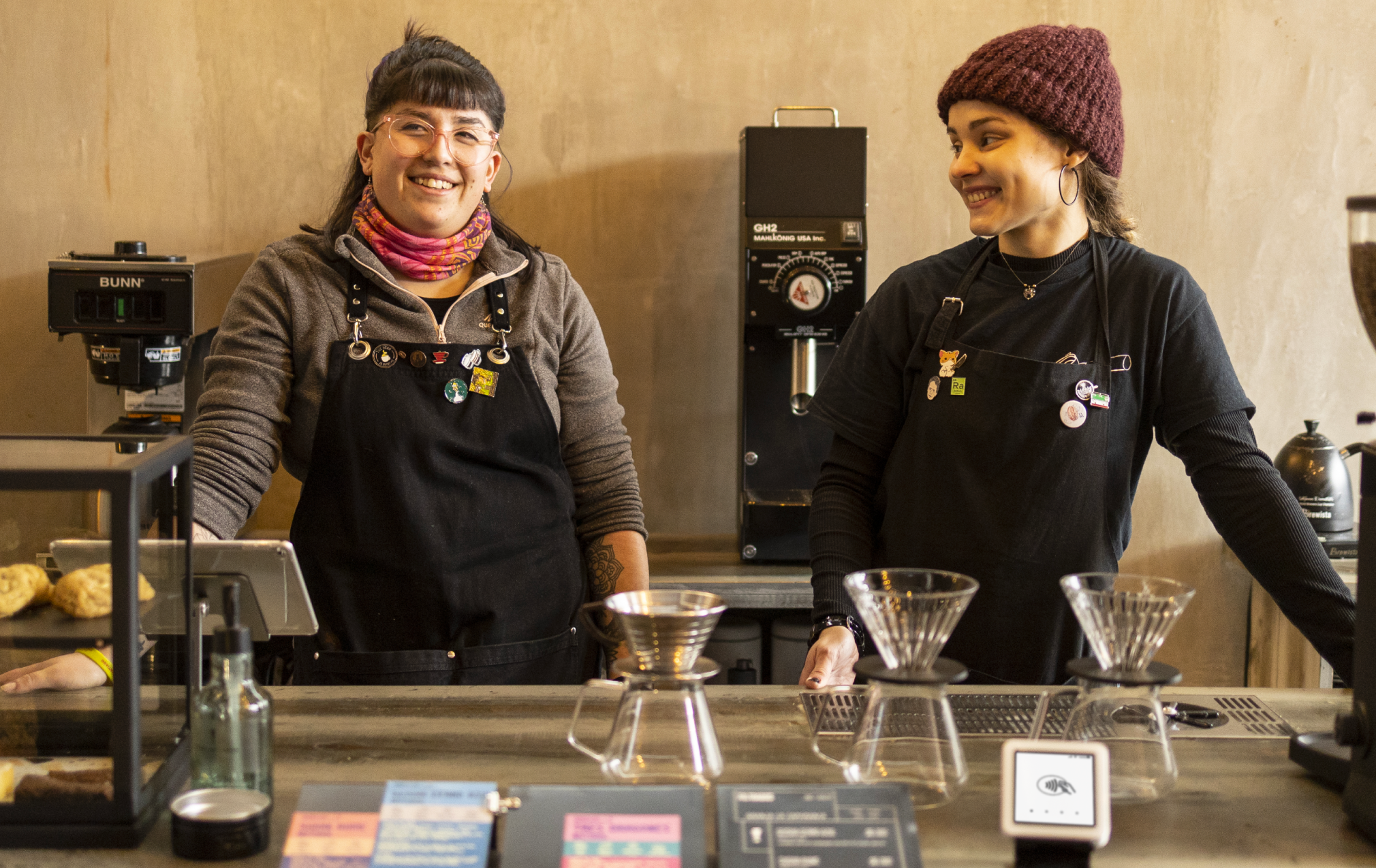 Két barista mosolyog a pult mögött, látható kávéfőző berendezéssel.