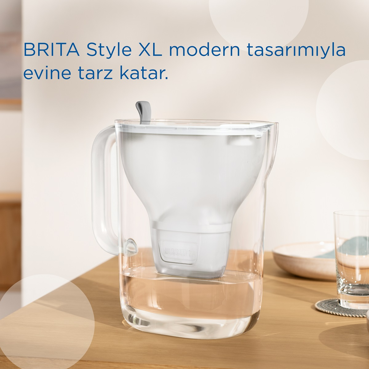 İçinde beyaz bir filtre bulunan şeffaf bir su sürahisi, ahşap bir masa üzerinde. Yanında bir bardak var.