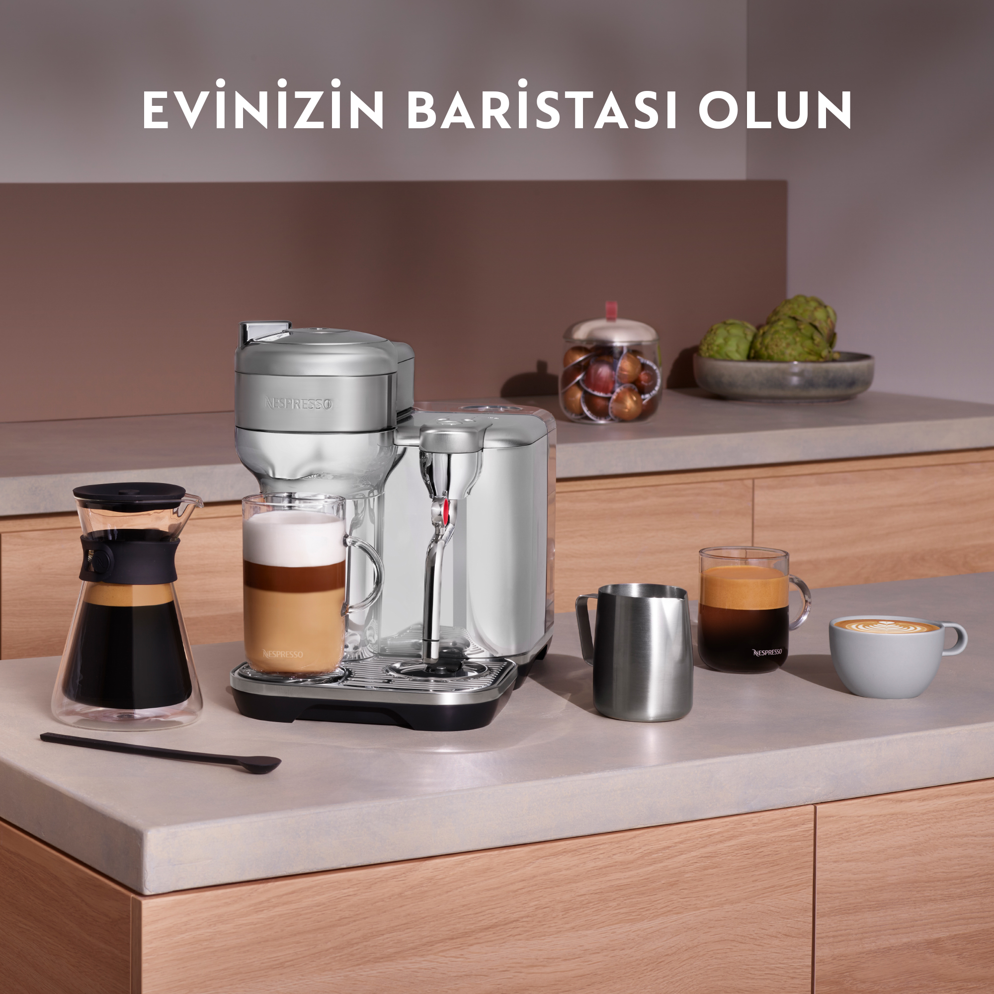 Latte'li kahve makinesi. Tezgahta fincanlar, sürahi ve bardaklar. Metin, 'Ev baristanız olun' diyor.