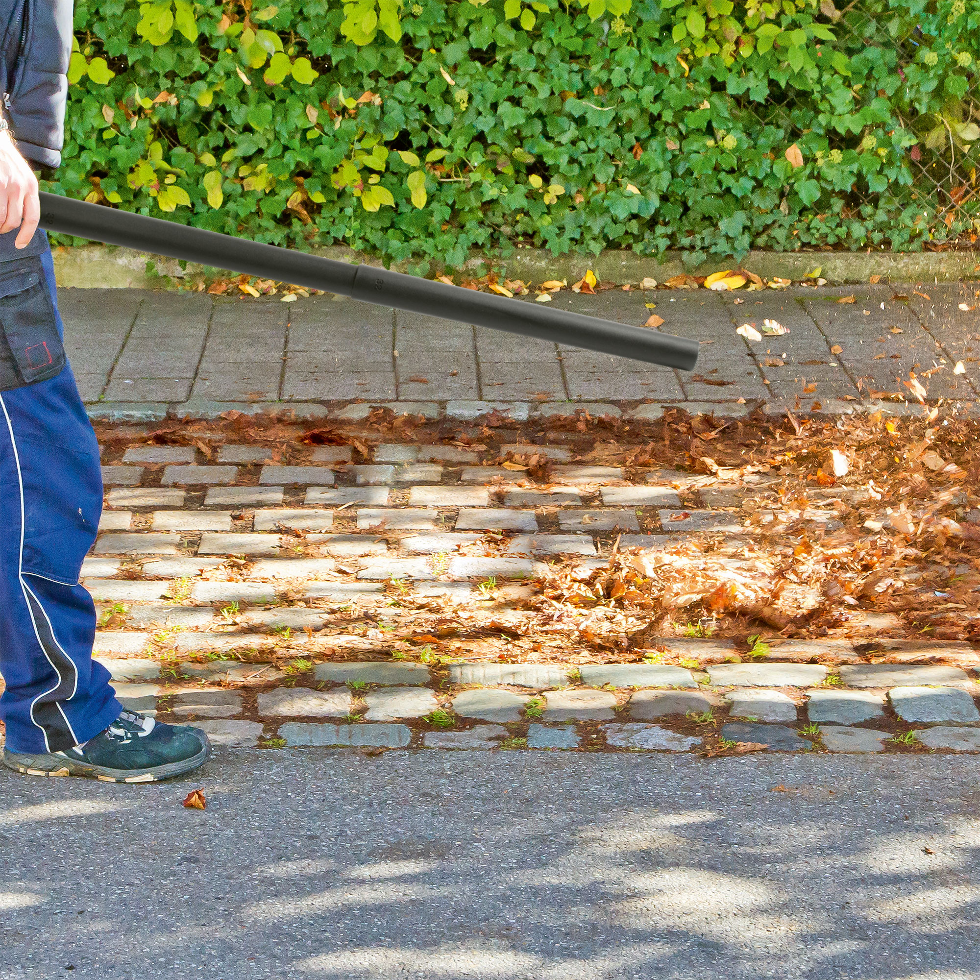 Person benutzt Laubbläser auf Pflasterfläche mit Blättern und Hecke.