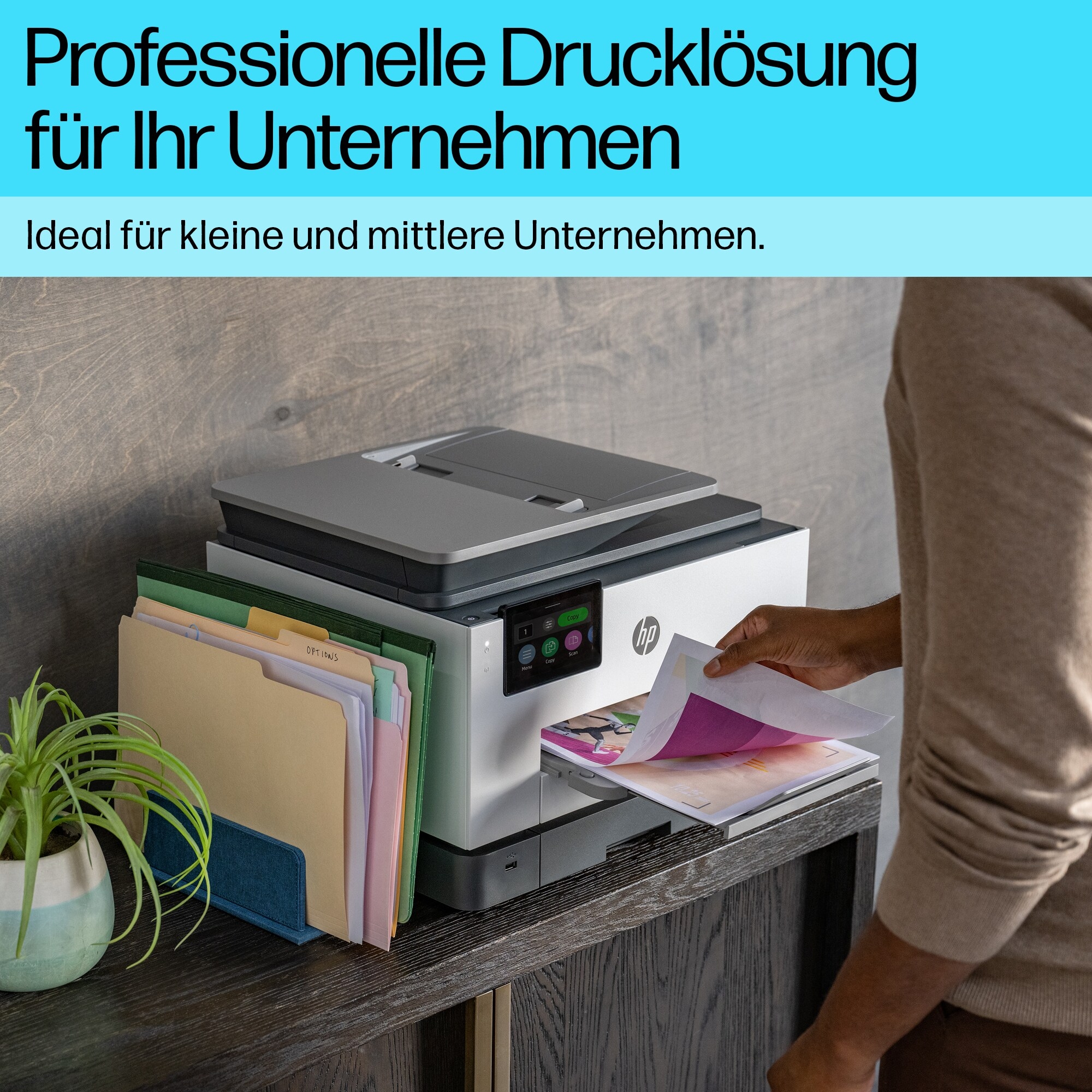 Ein Mann nimmt Papier aus einem Drucker. Links befinden sich eine Pflanze und Akten.