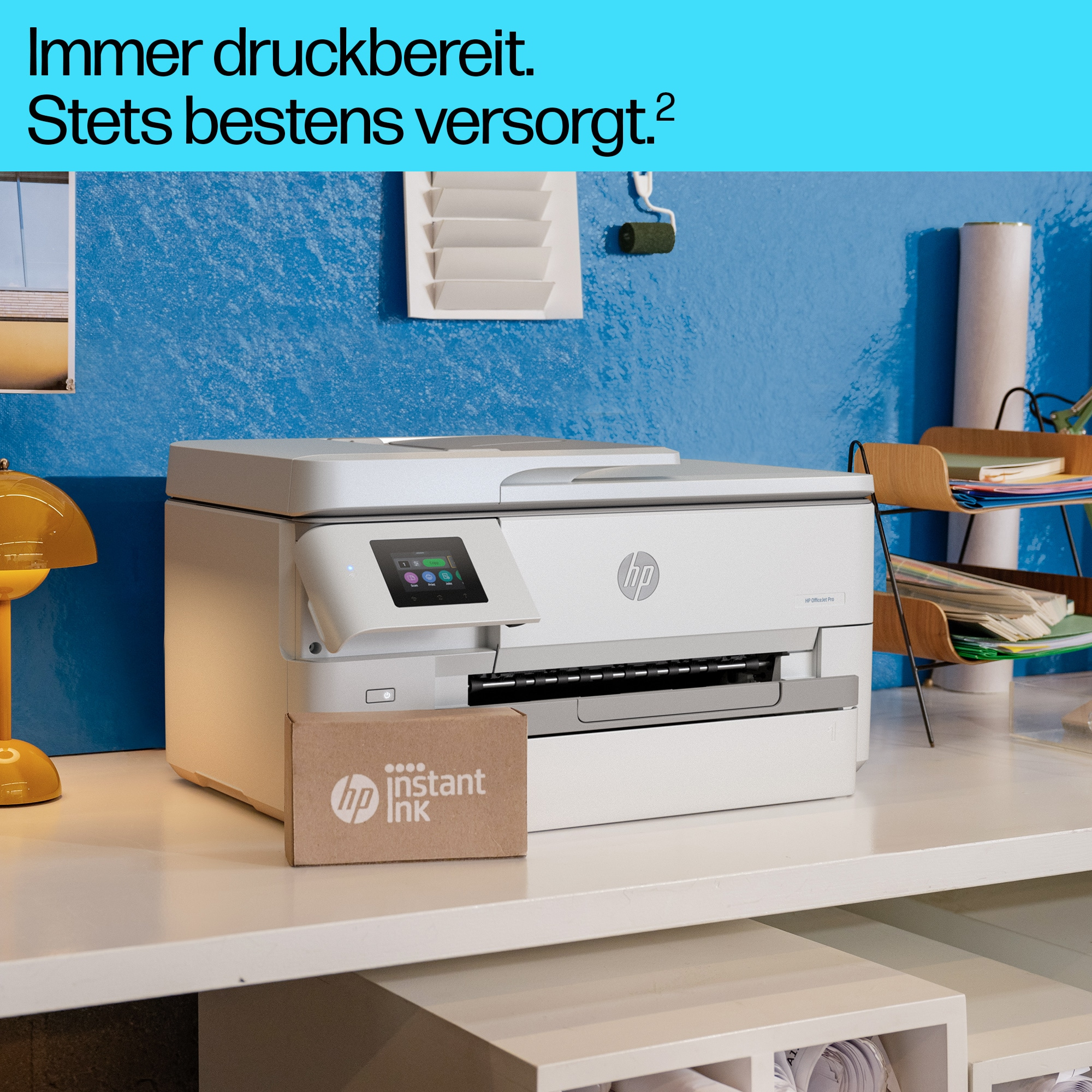 Weißer Drucker auf einem Schreibtisch. Karton mit dem Text 'HP Instant Ink'. Blauer Wandhintergrund.