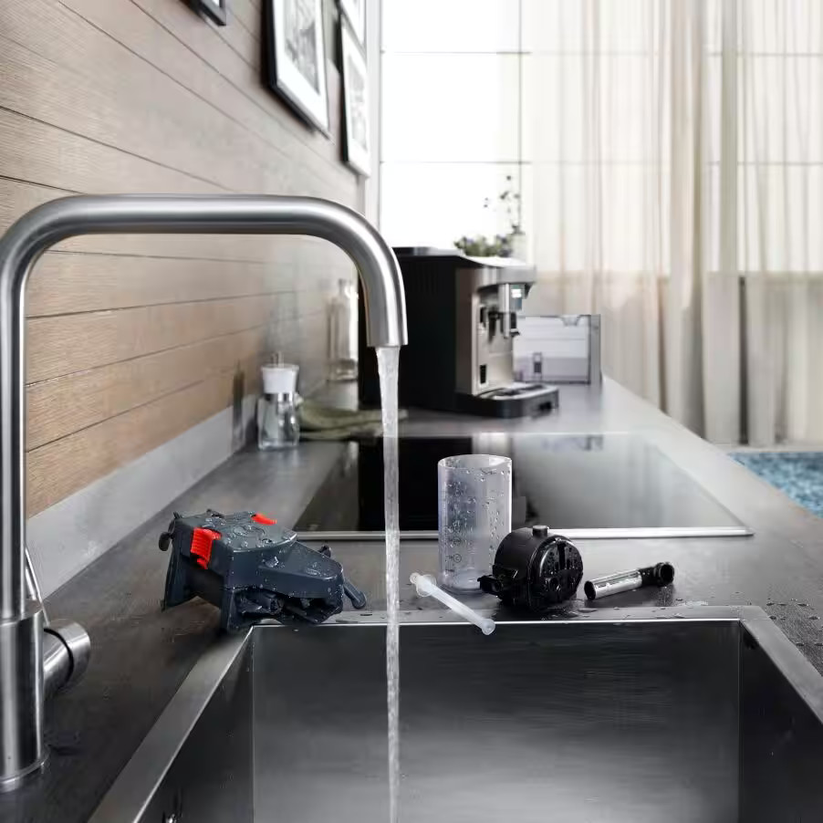 Zlew kuchenny z bieżącą wodą i sprzętem do czyszczenia. W tle widoczny jest ekspres do kawy.