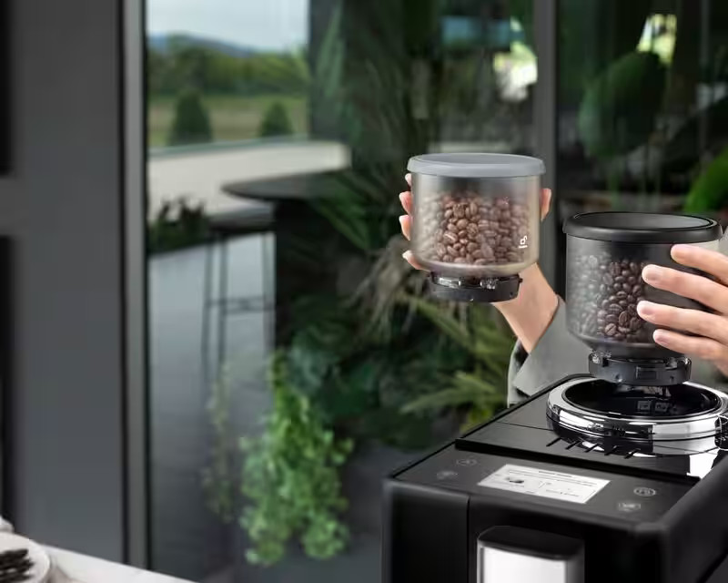 Dłonie trzymające dwa przezroczyste pojemniki wypełnione ziarnami kawy nad ekspresem do kawy. Tło z zielenią.