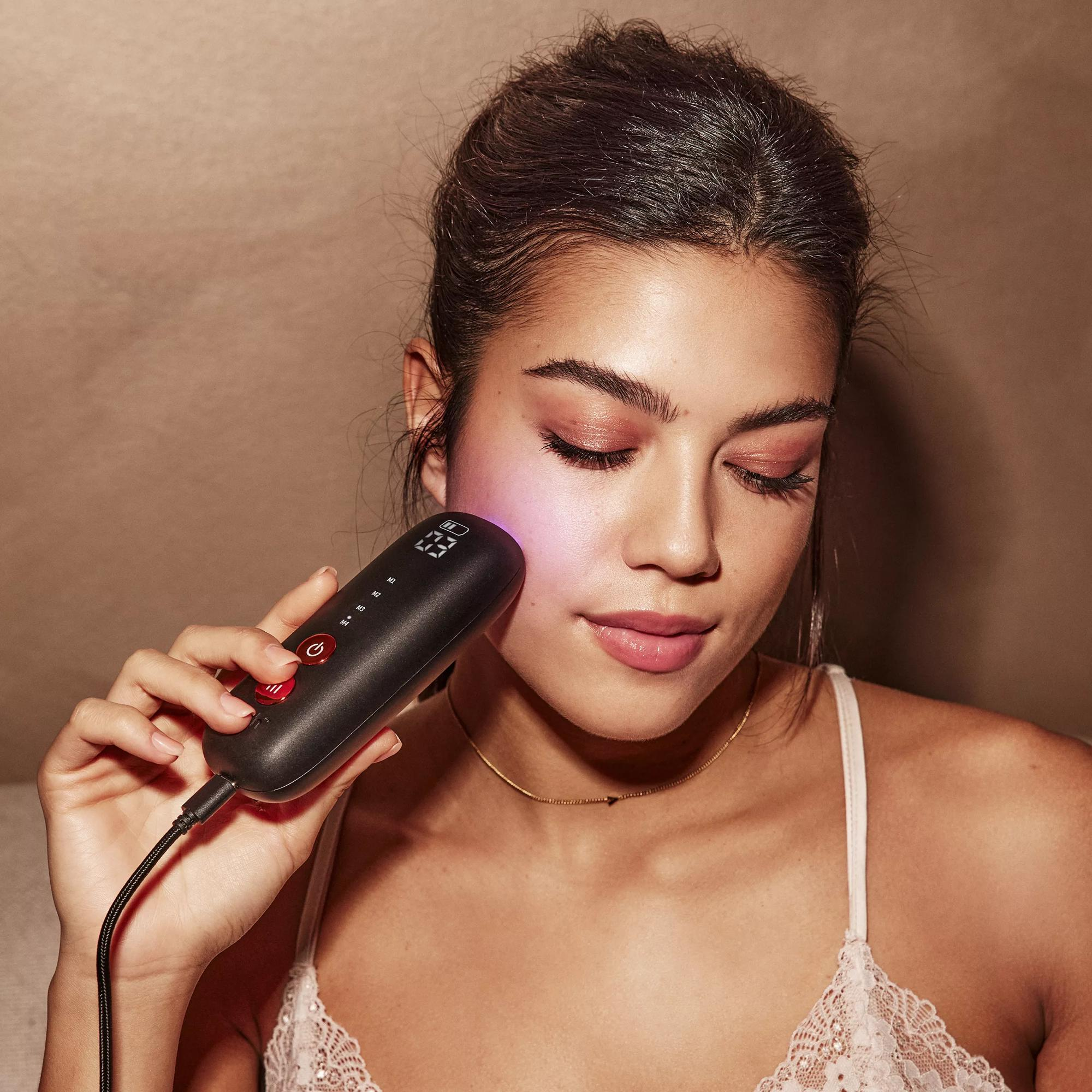 Kobieta używa czarnego urządzenia do pielęgnacji skóry na twarzy. Urządzenie ma czerwony przycisk.