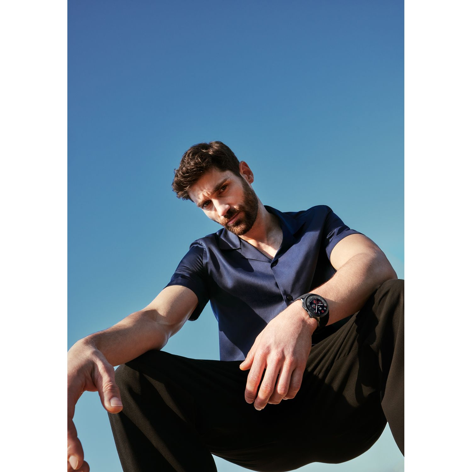 Homme en chemise bleue regardant vers le bas, portant une montre et un pantalon noir, sur fond de ciel bleu.