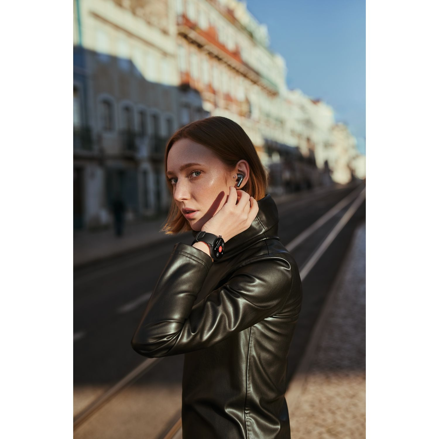 Femme portant des écouteurs et une veste noire, debout dans une rue avec des bâtiments.