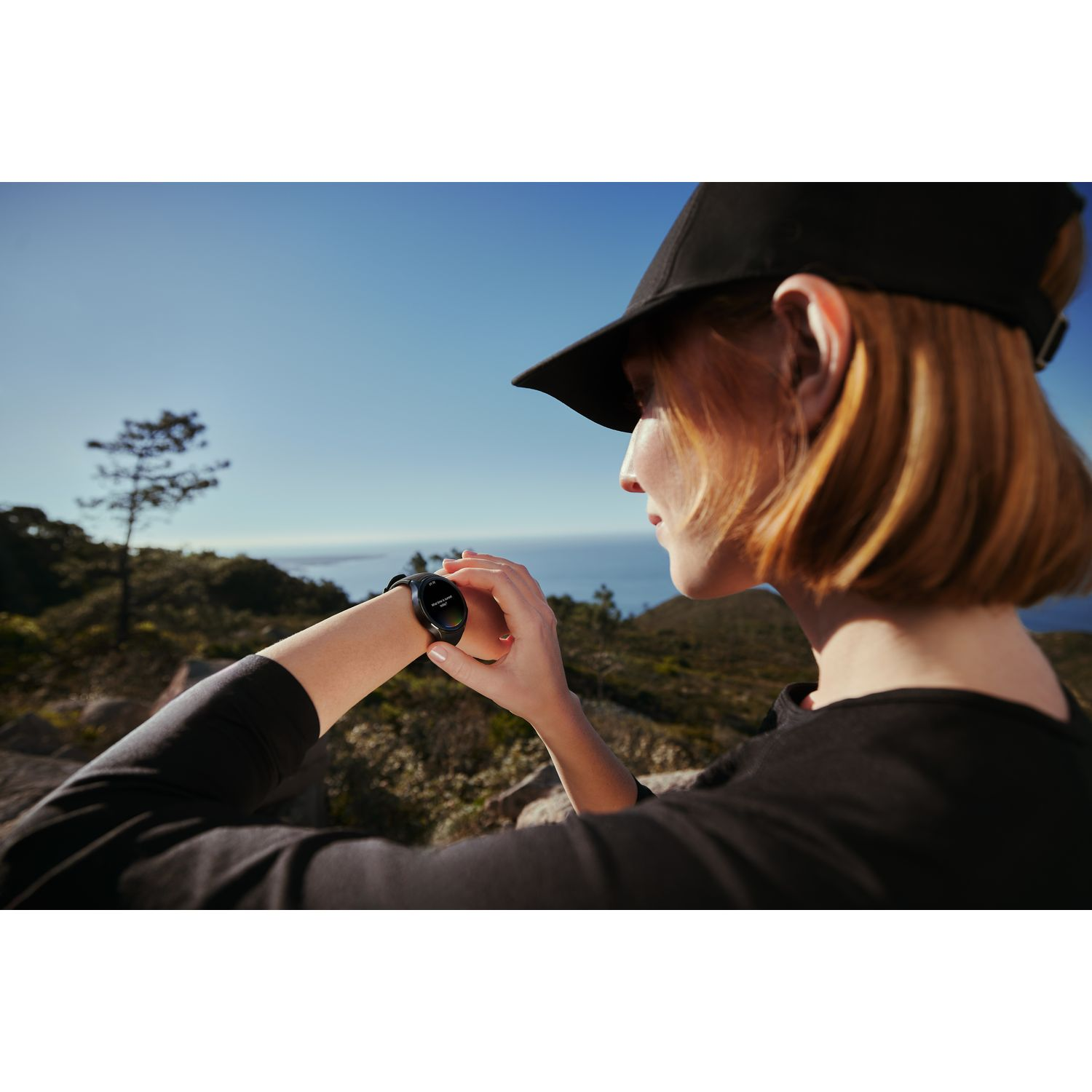 Une femme en casquette et chemise noires regarde une montre connectée à son poignet sur fond de ciel bleu.