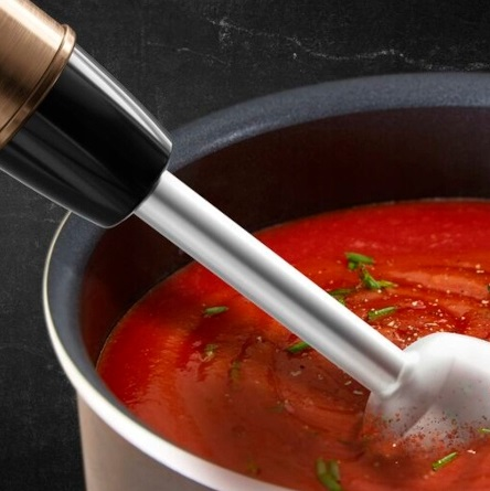 Blender ręczny w garnku z czerwonym sosem. Blender jest srebrny i czarny.