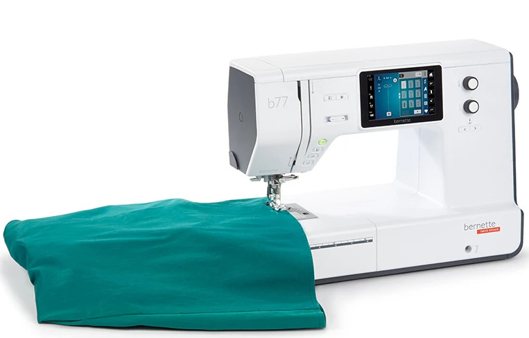 Biała maszyna do szycia z zieloną tkaniną pod spodem, na białym tle.