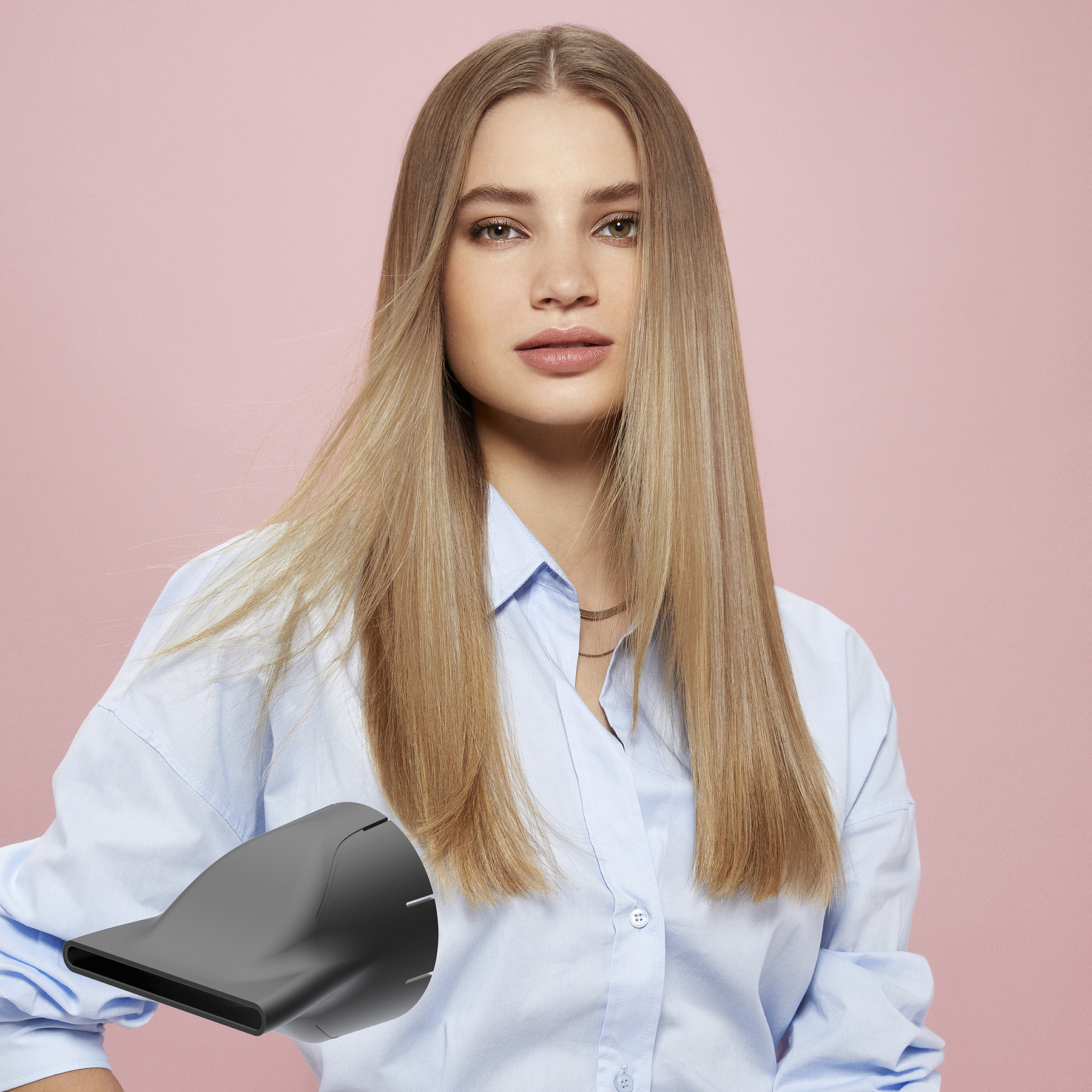 Une femme aux cheveux blonds utilise un sèche-cheveux noir sur un fond rose.