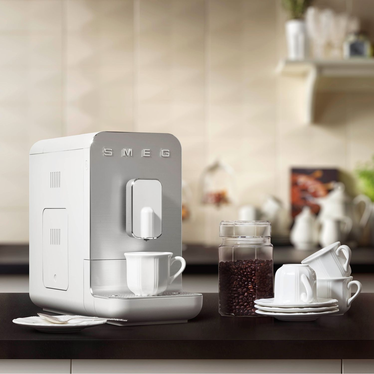 A white SMEG coffee machine, coffee beans, cups, and saucers on a dark countertop.