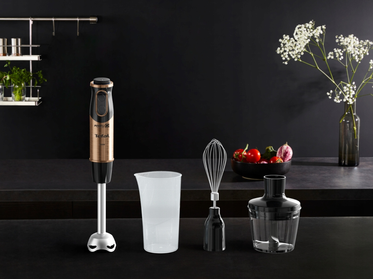 Blat kuchenny z czarnym blenderem, miarką, trzepaczką oraz miską warzyw i kwiatów.