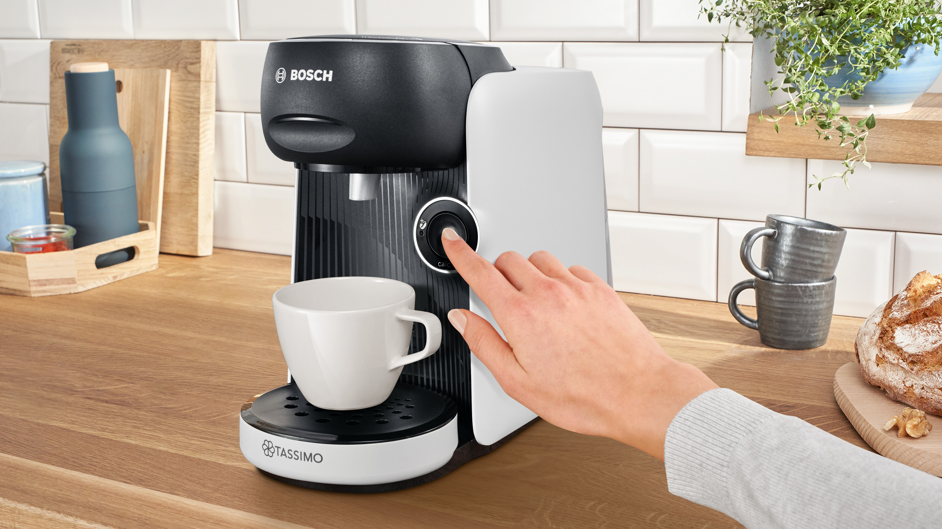 Hand einer Person, die einen Knopf an einer schwarz-weißen Kaffeemaschine drückt. Weiße Tasse auf einem Tablett. Nahaufnahme.