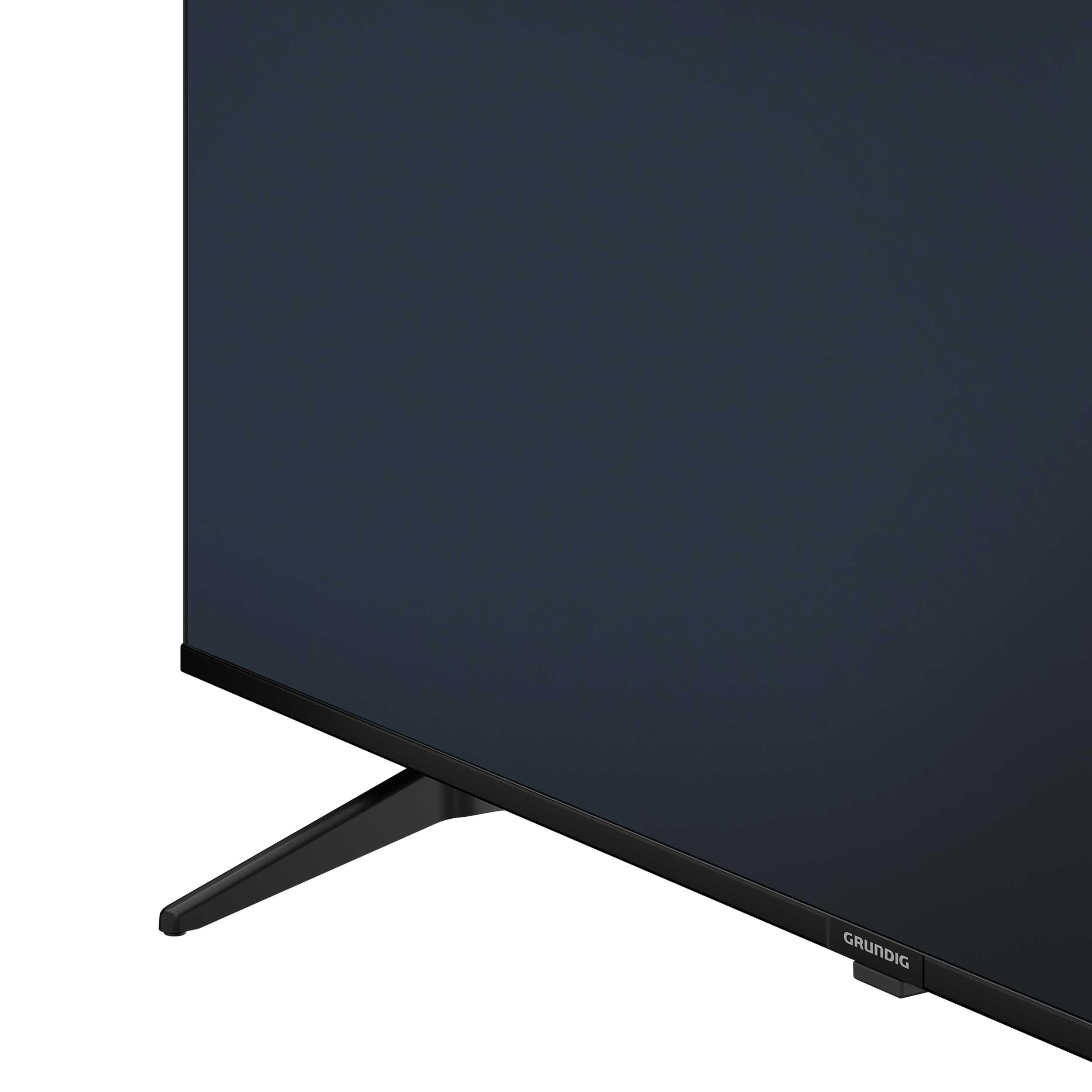Ecke eines schwarzen Grundig-Fernsehers mit schwarzem Ständer auf weißem Hintergrund.