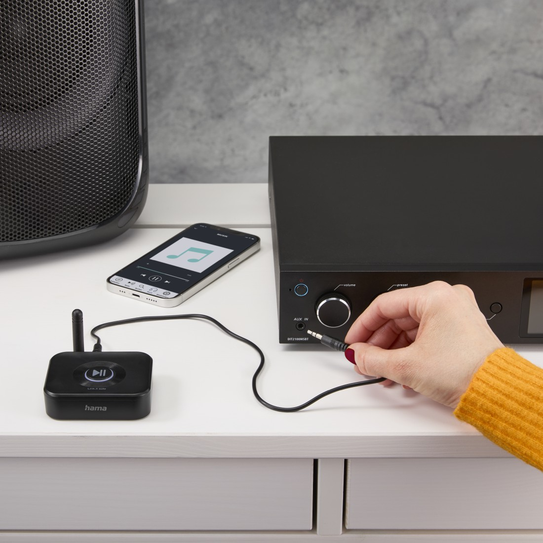 Eine Hand steckt ein schwarzes Audiokabel in ein schwarzes Gerät auf einem weißen Tisch, mit Telefon und Lautsprecher.