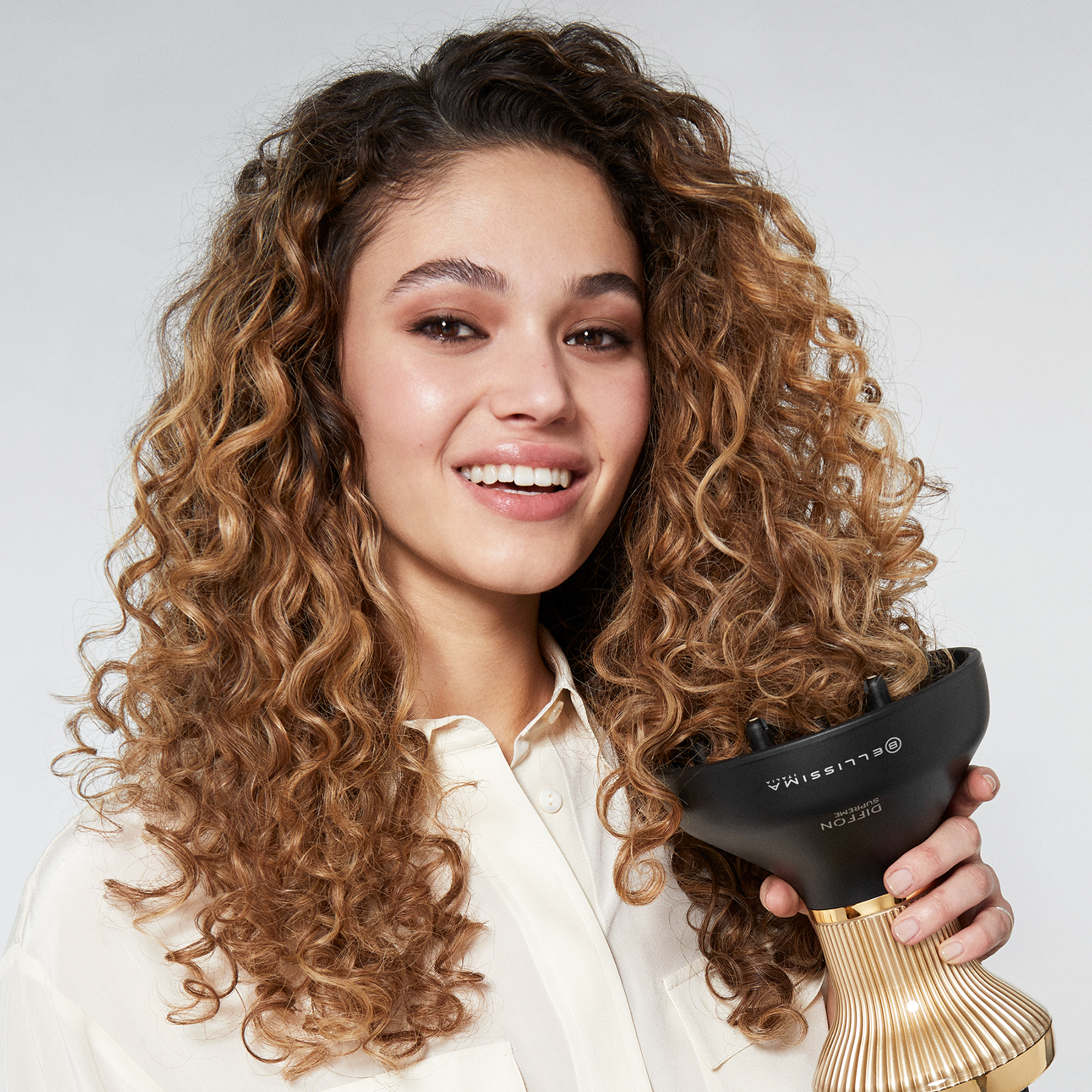 Femme aux cheveux bouclés souriant, tenant un diffuseur de cheveux noir et doré.