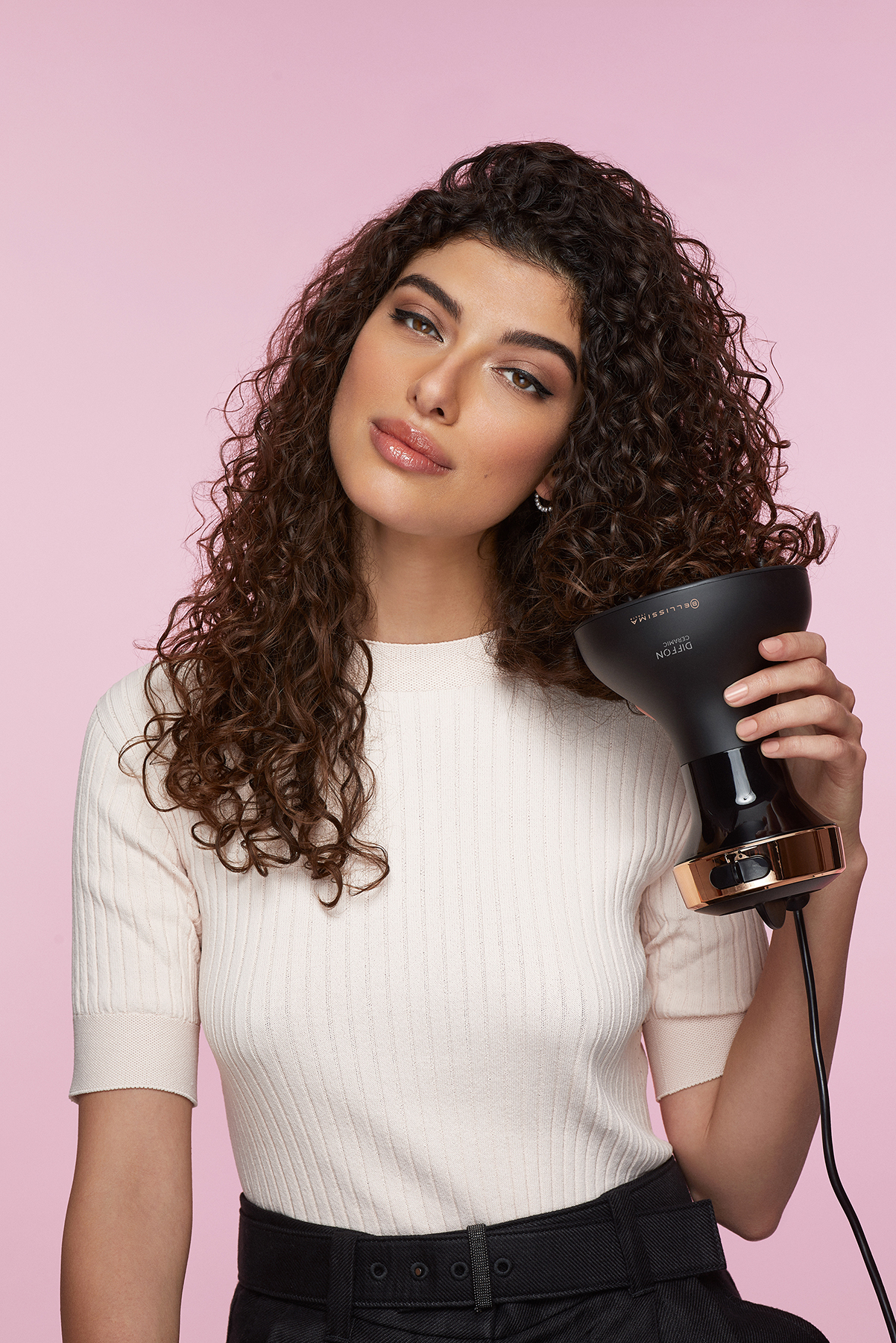 Une femme tient un diffuseur de cheveux noir. Elle porte une chemise rose clair et un pantalon noir.