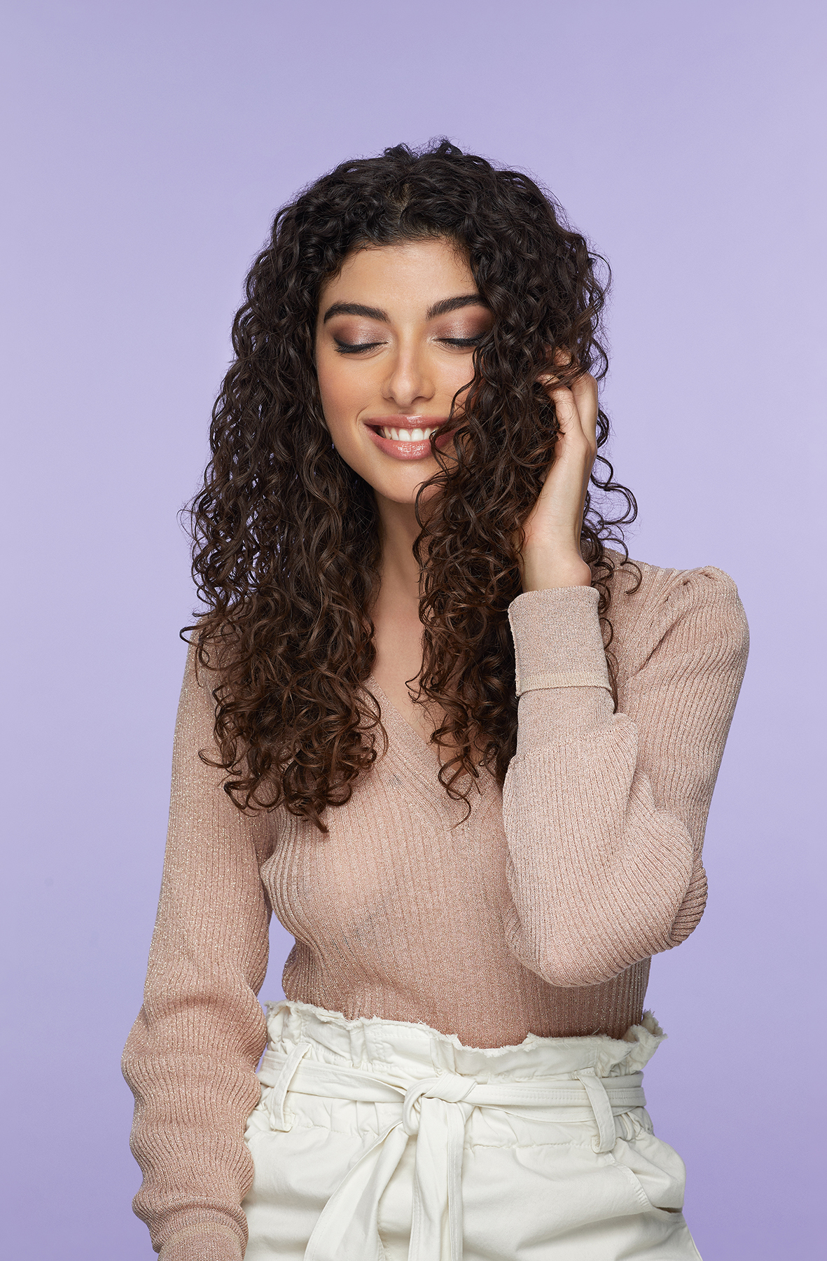 Une femme aux cheveux bouclés dans un pull rose clair. Elle sourit et se touche les cheveux.
