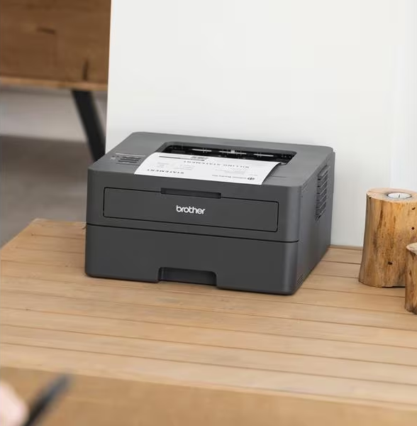 Une imprimante Brother grise imprimant un document sur une table en bois, avec un mur blanc.