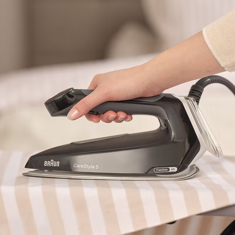Hand bügelt Stoff mit einem schwarz-grauen Bügeleisen. Das Bügeleisen hat ein Braun-Logo.