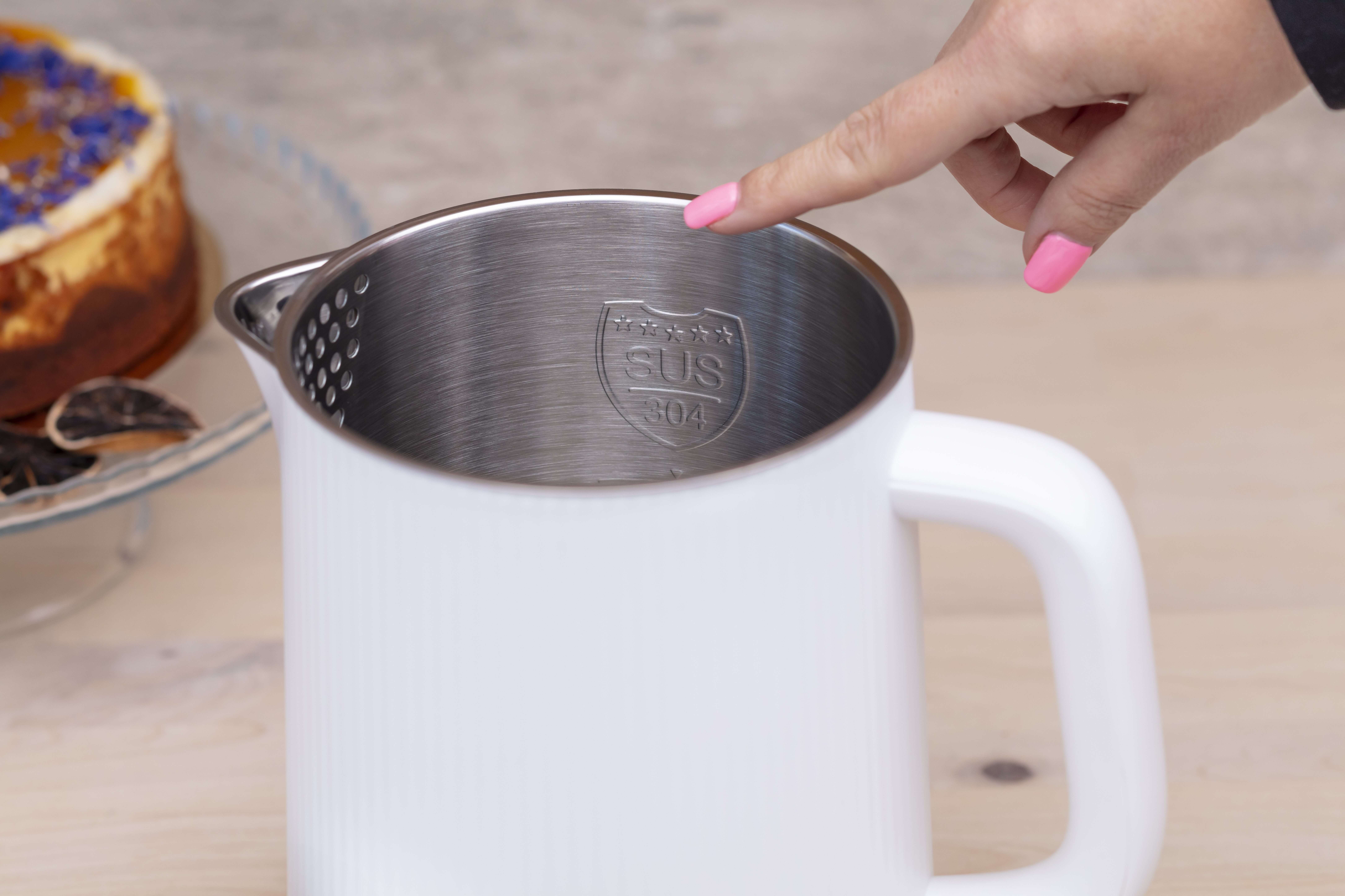 Ręka wskazuje na wnętrze białego czajnika. Ciasto w tle.