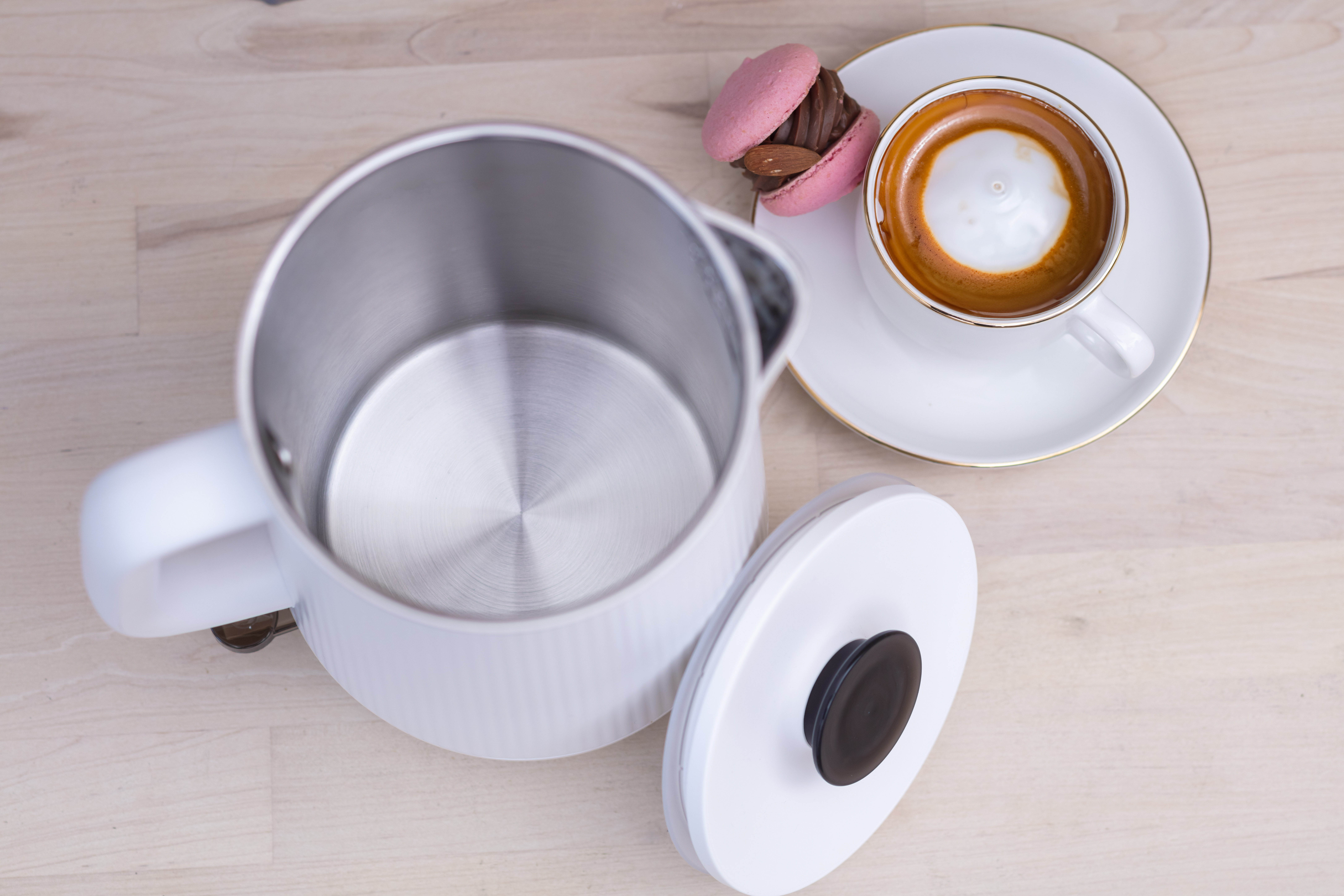 Biały czajnik, filiżanka kawy i różowy makaronik na drewnianym stole.