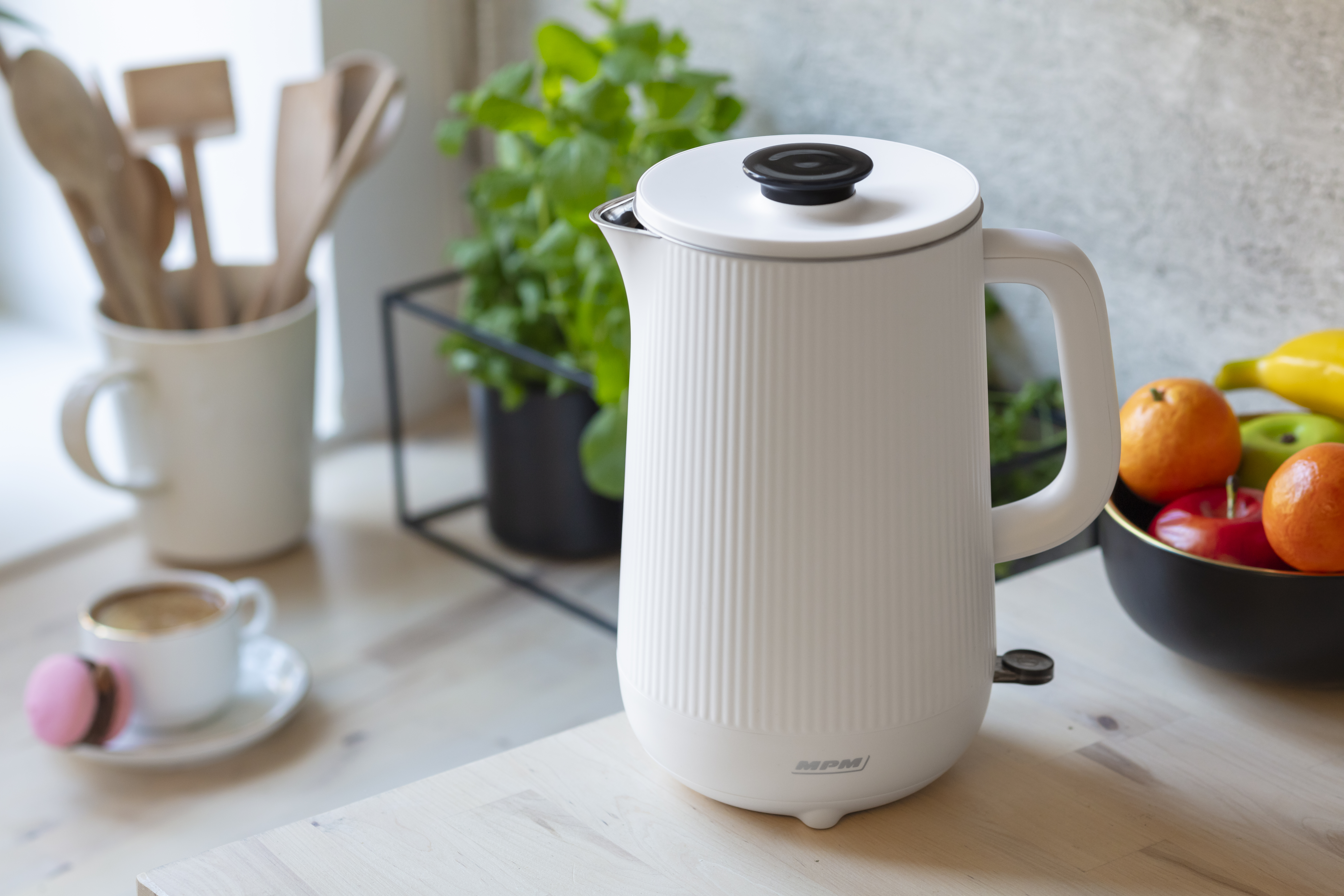 Biały czajnik elektryczny z czarnym wieczkiem. Na drewnianym stole z filiżanką, deserem i owocami.