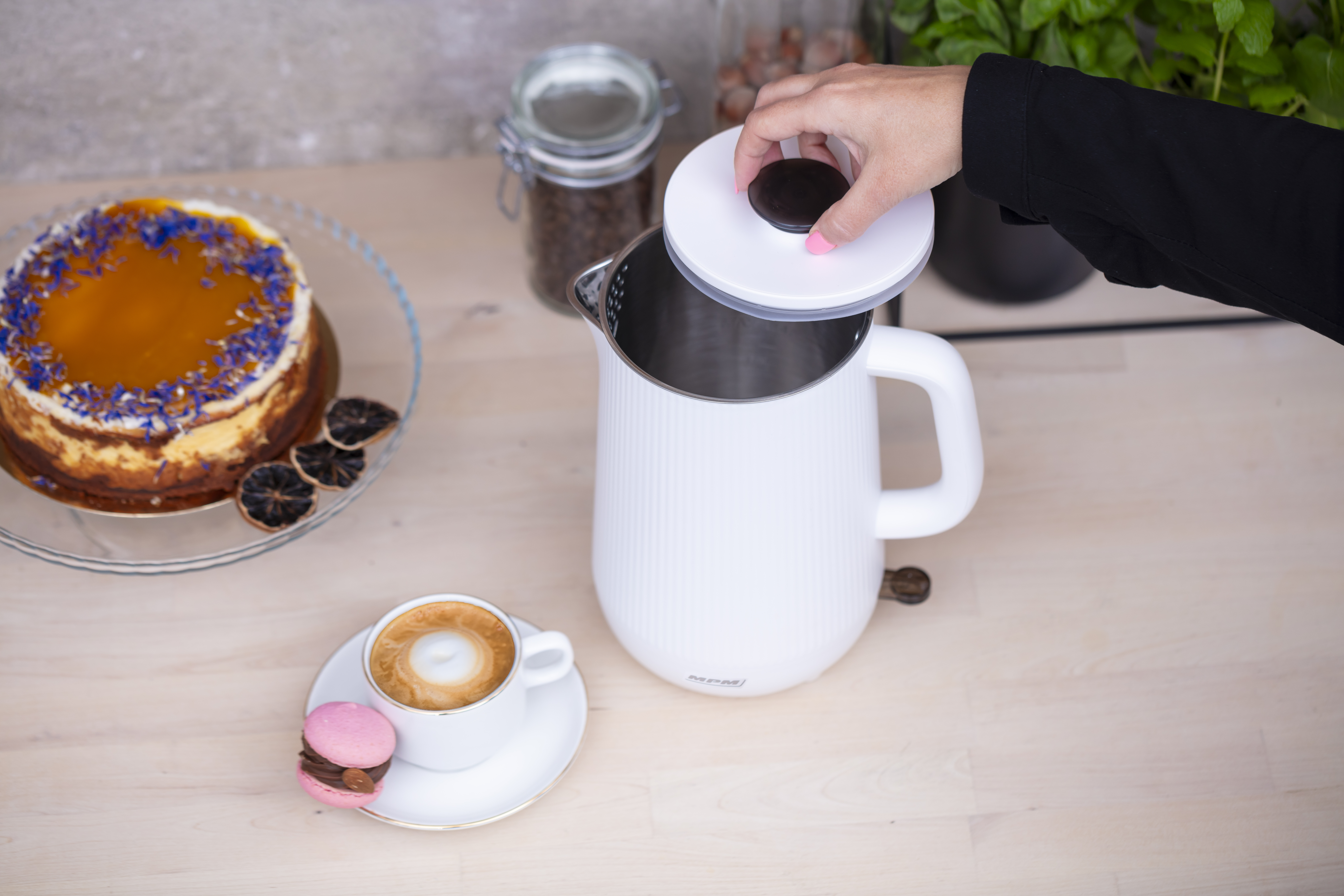 Osoba otwiera biały czajnik elektryczny. Na stole filiżanka kawy i ciasto.