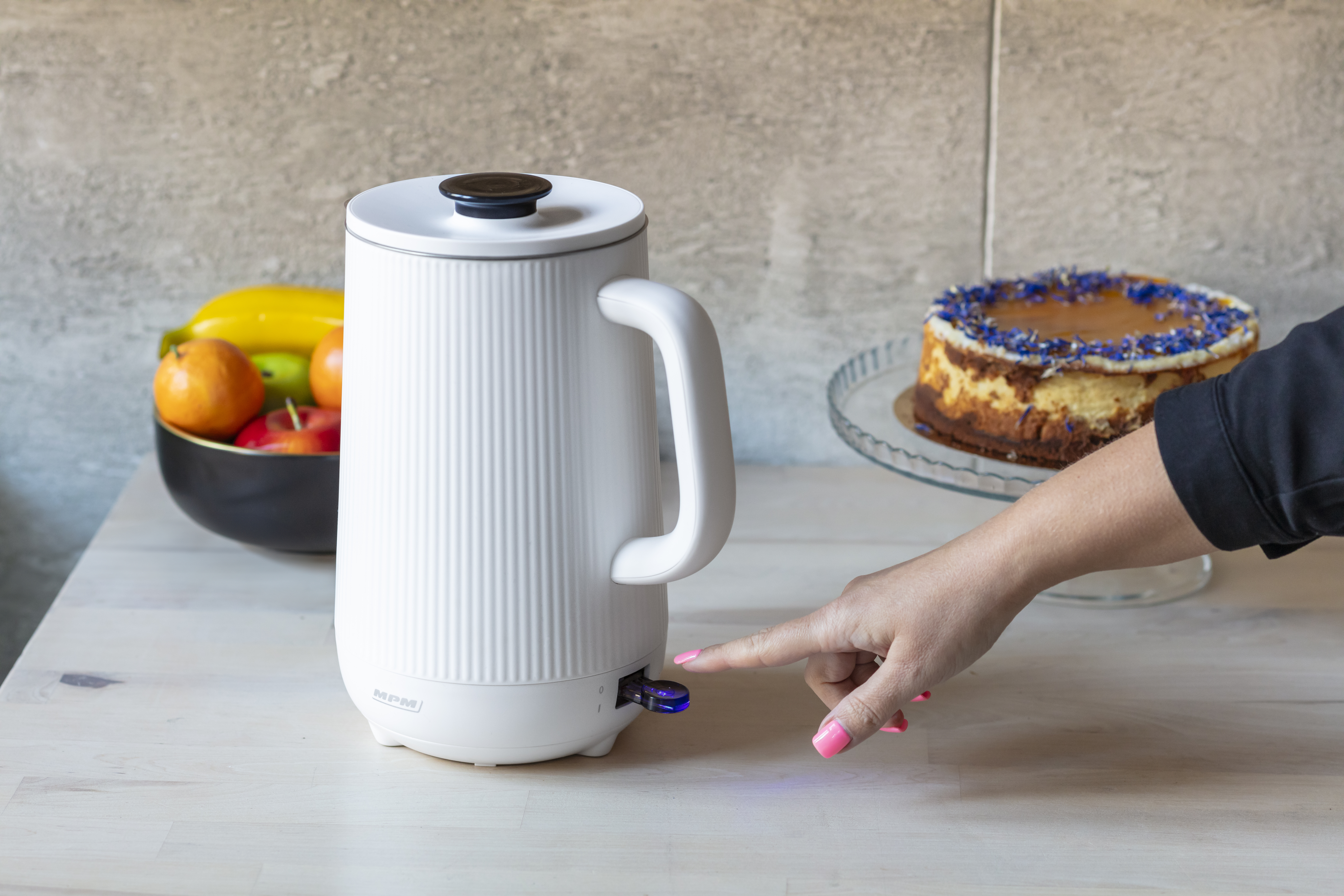 Biały czajnik elektryczny z ręką naciskającą przycisk zasilania i ciastem w tle.