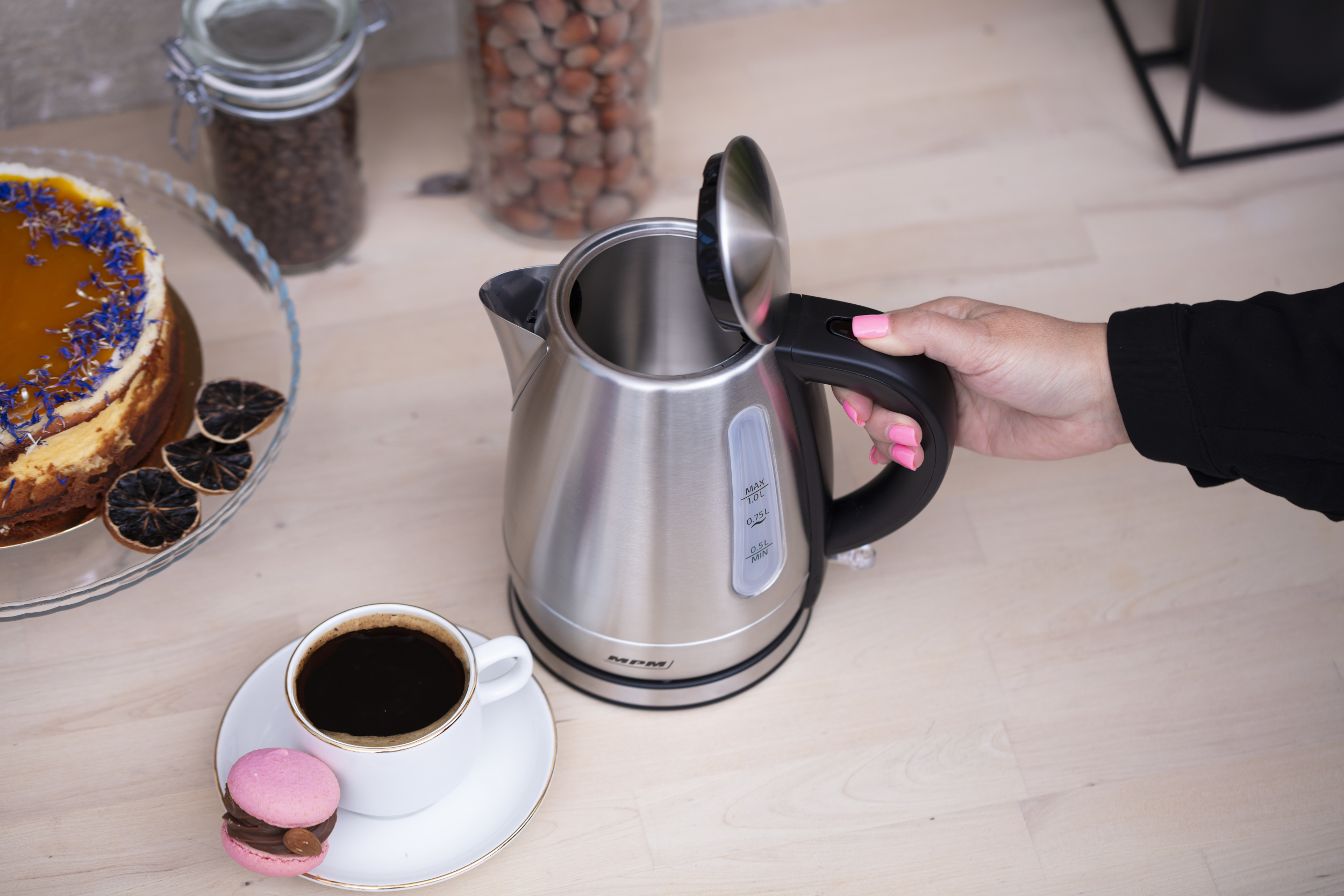 Czajnik ze stali nierdzewnej z czarnym uchwytem. Obok filiżanka kawy i ciastko.