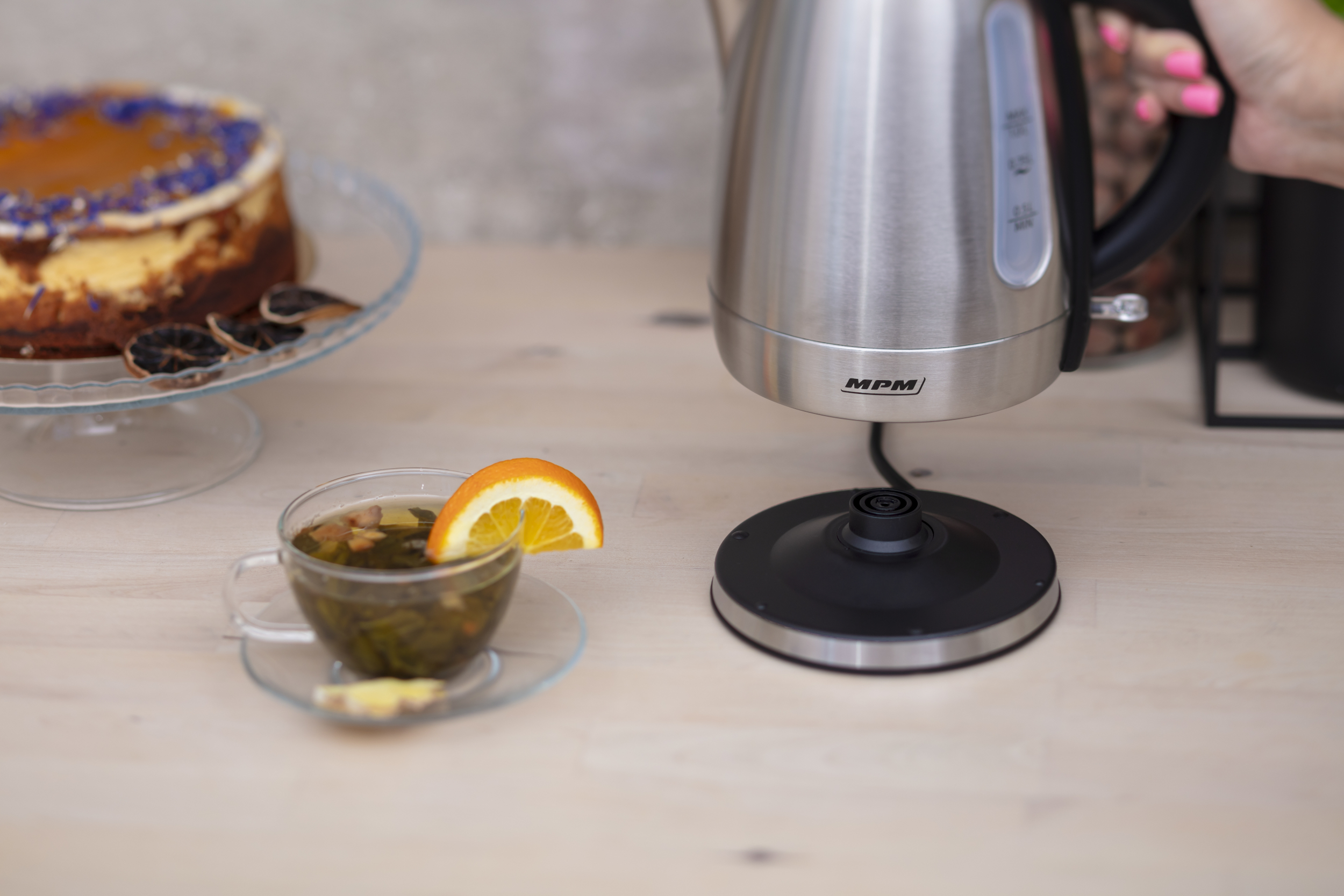 Filiżanka herbaty ze skórką pomarańczy. Czajnik na podstawce. Ciasto na szklanym talerzu.