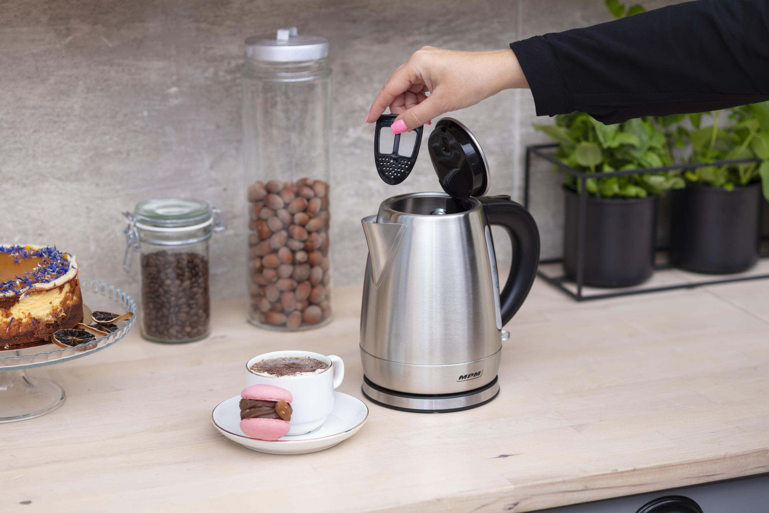 Kobieta umieszcza filtr w srebrnym czajniku. Kawa, makaronik, orzechy i rośliny.