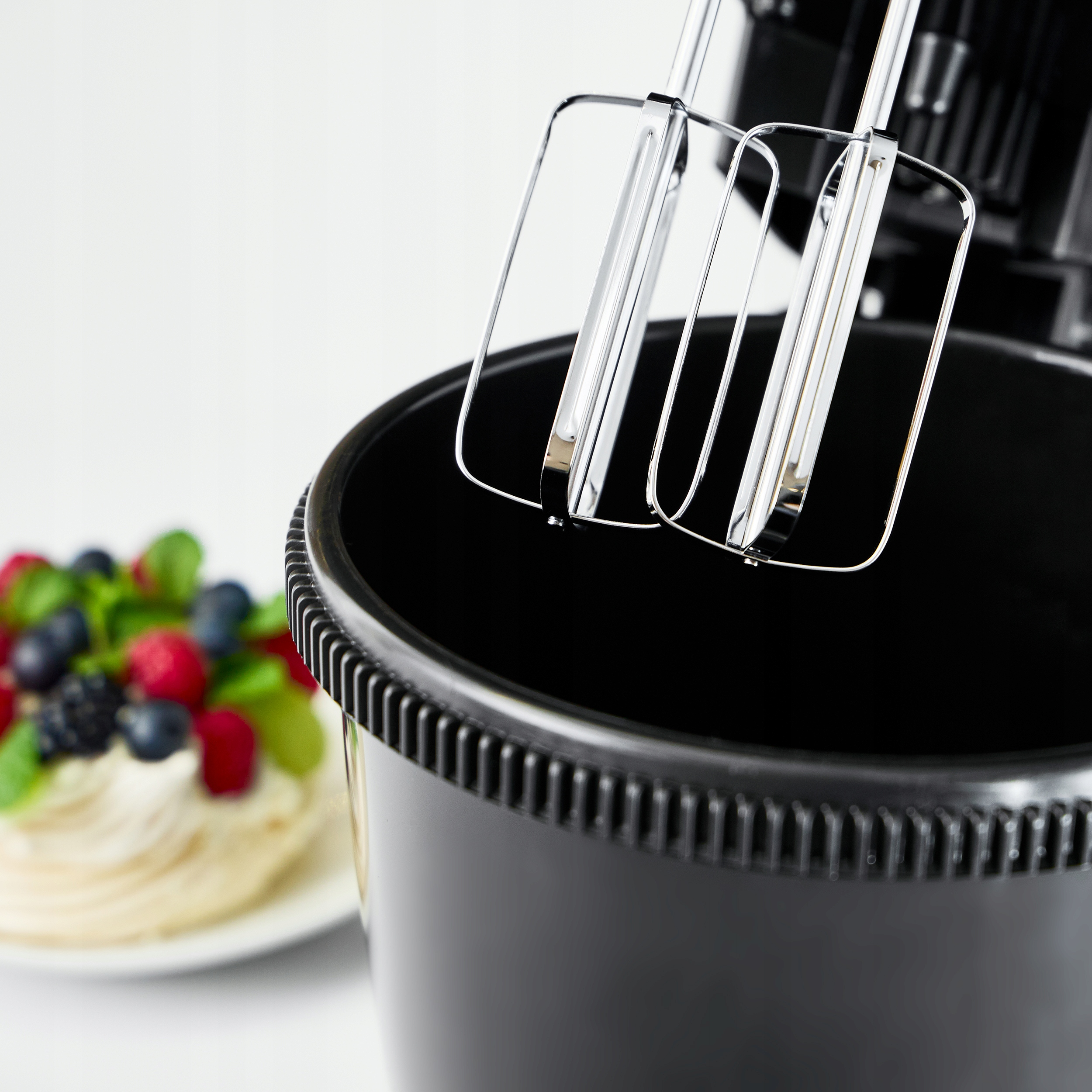 Zbliżenie czarnej miski miksera z końcówkami do ubijania i talerzem z deserem.