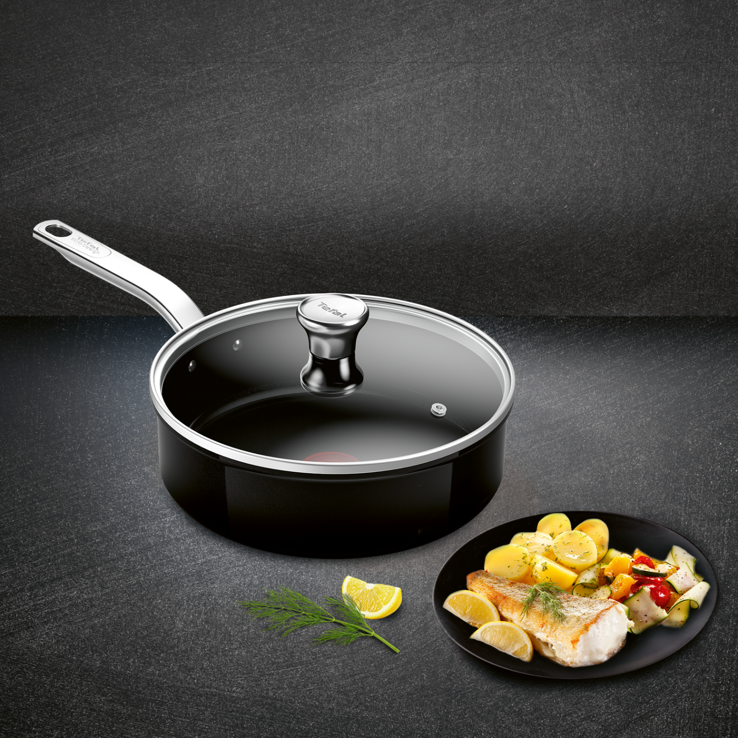 Black frying pan with a glass lid and food on a plate, with a lemon slice.