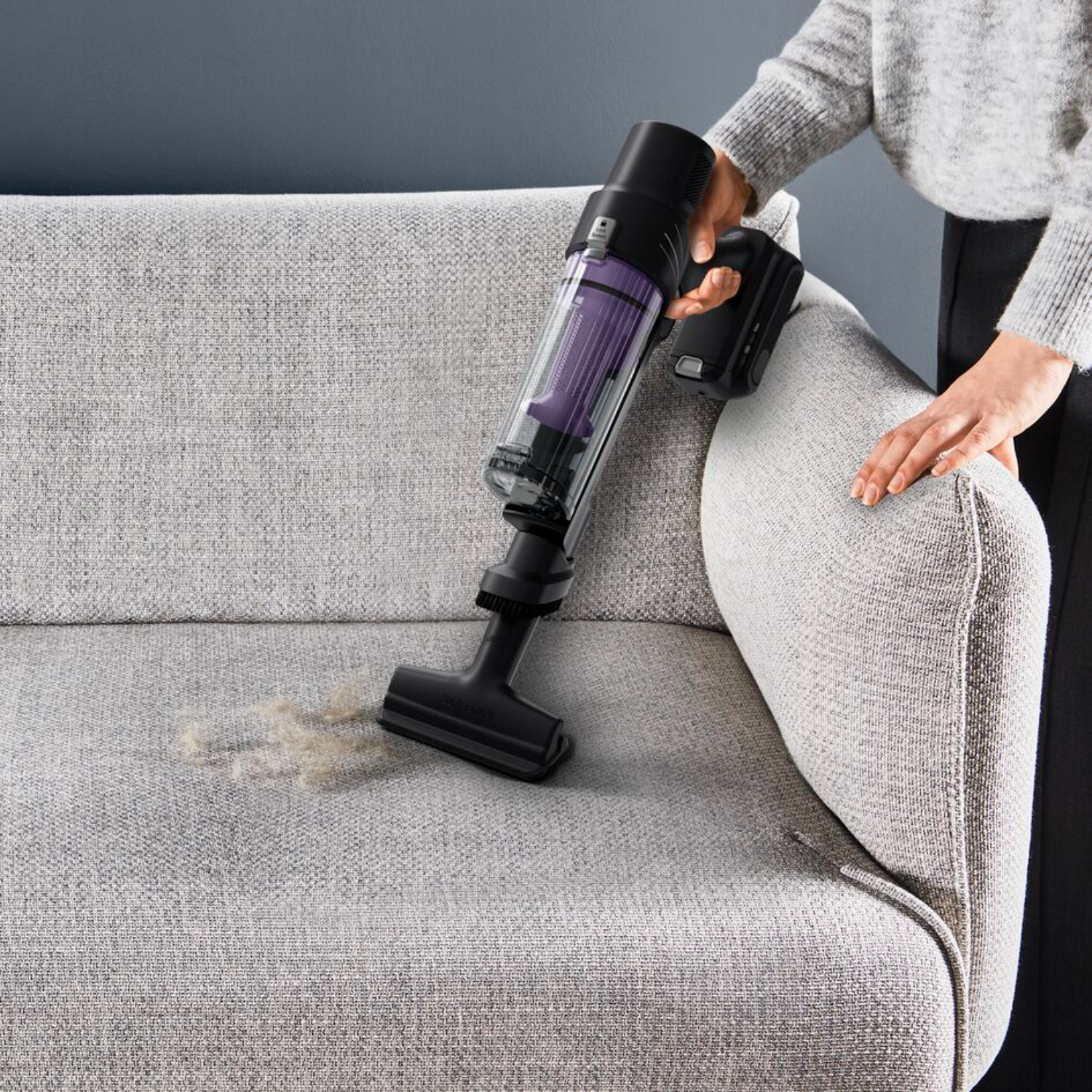A person vacuums a gray sofa with a black and purple cordless vacuum cleaner.