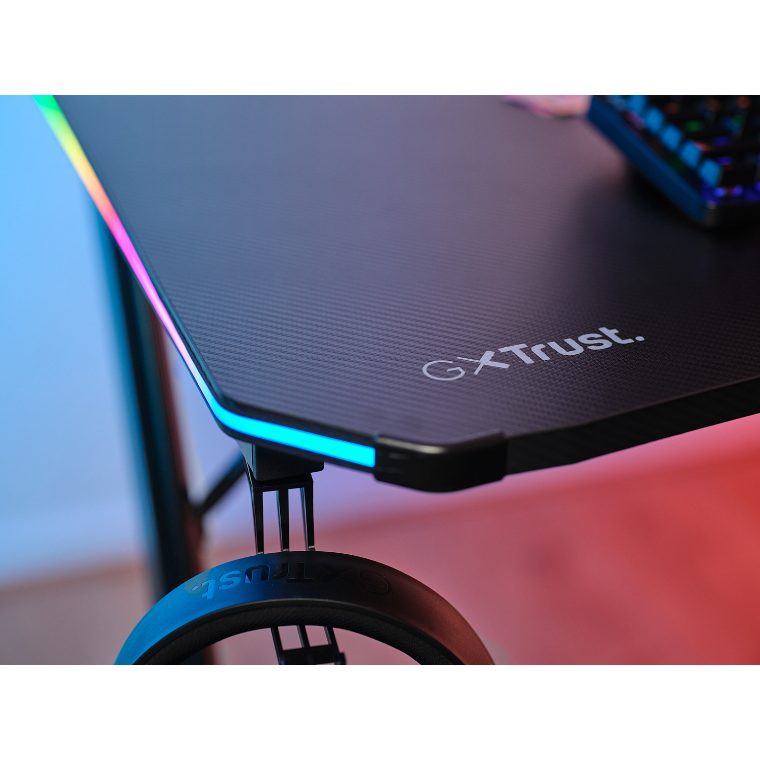 Primo piano di un angolo di scrivania da gaming nera con testo 'GXTtrust' e supporto cuffie.