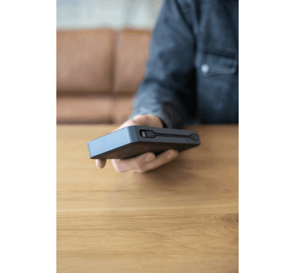 Una persona sosteniendo un banco de energía rectangular gris oscuro sobre una mesa de madera.