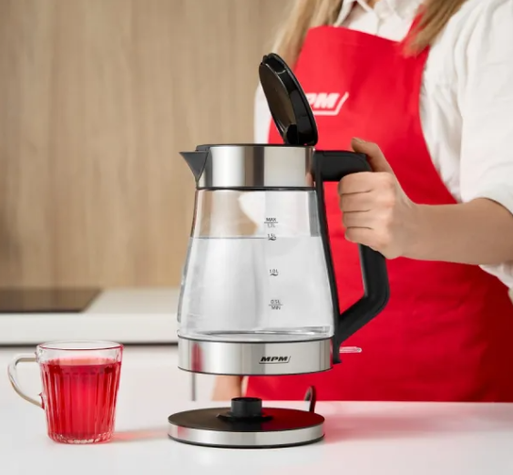 Kobieta trzymająca przezroczysty szklany czajnik elektryczny nad białym blatem, ze szklanką soku.